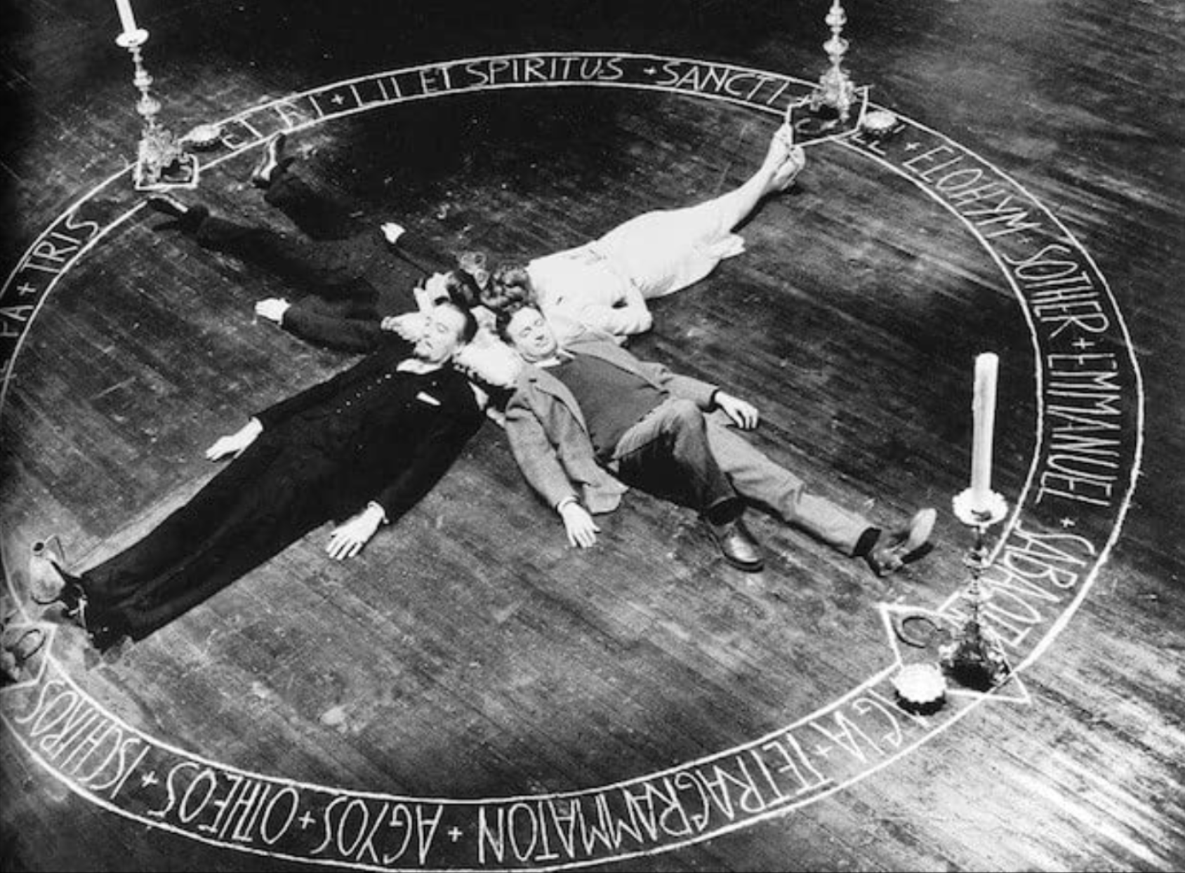 Trois personnes allongées sur un plancher en bois, formant un cercle autour d'une inscription circulaire. L'inscription est en latin et quelques mots sont visibles, tels que 'SPIRITUS', 'SANCTI', 'YOUTUBE'. La scène semble être une reconstitution ou une œuvre artistique avec des éléments symboliques.