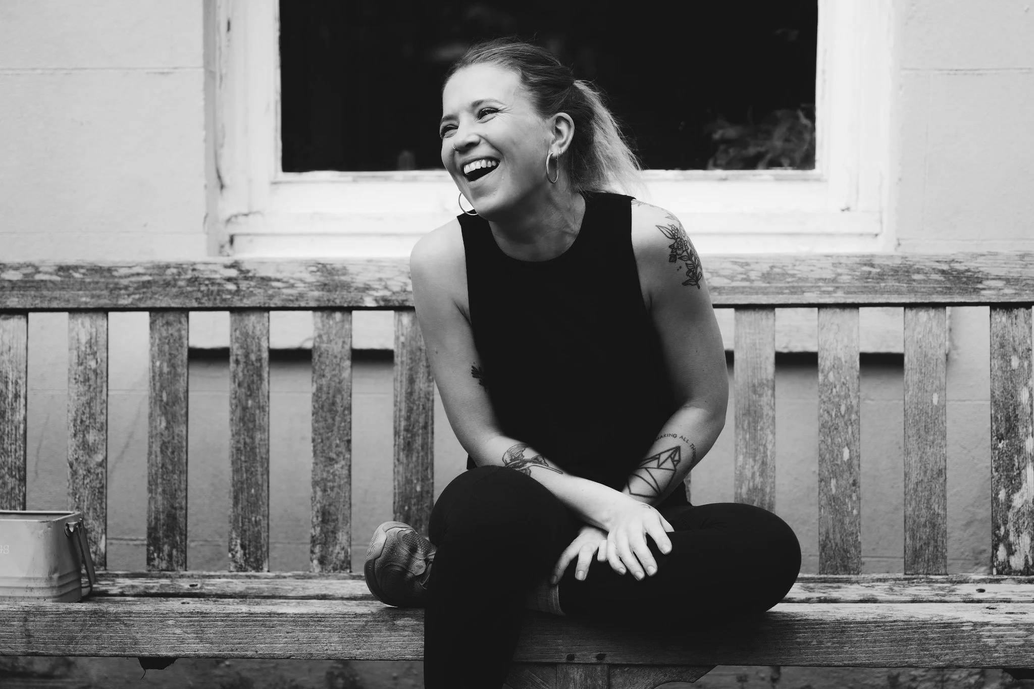 A woman with tattoos and hoop earrings is sitting cross-legged on a wooden bench, laughing and leaning slightly forward, with a window behind her.