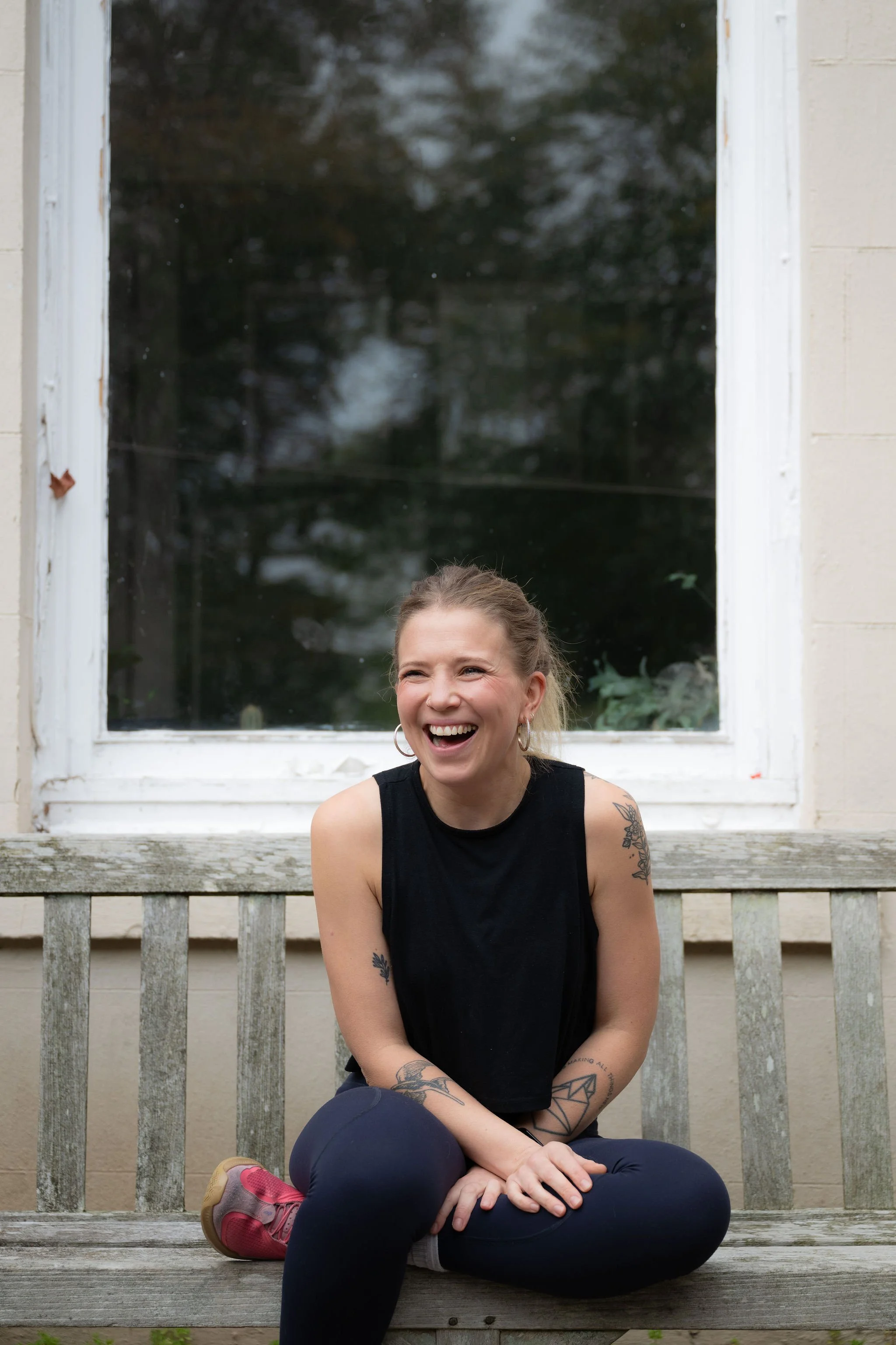 A woman with tattoos, wearing a black sleeveless top and black leggings, sitting on a worn wooden bench in front of a large window, laughing.