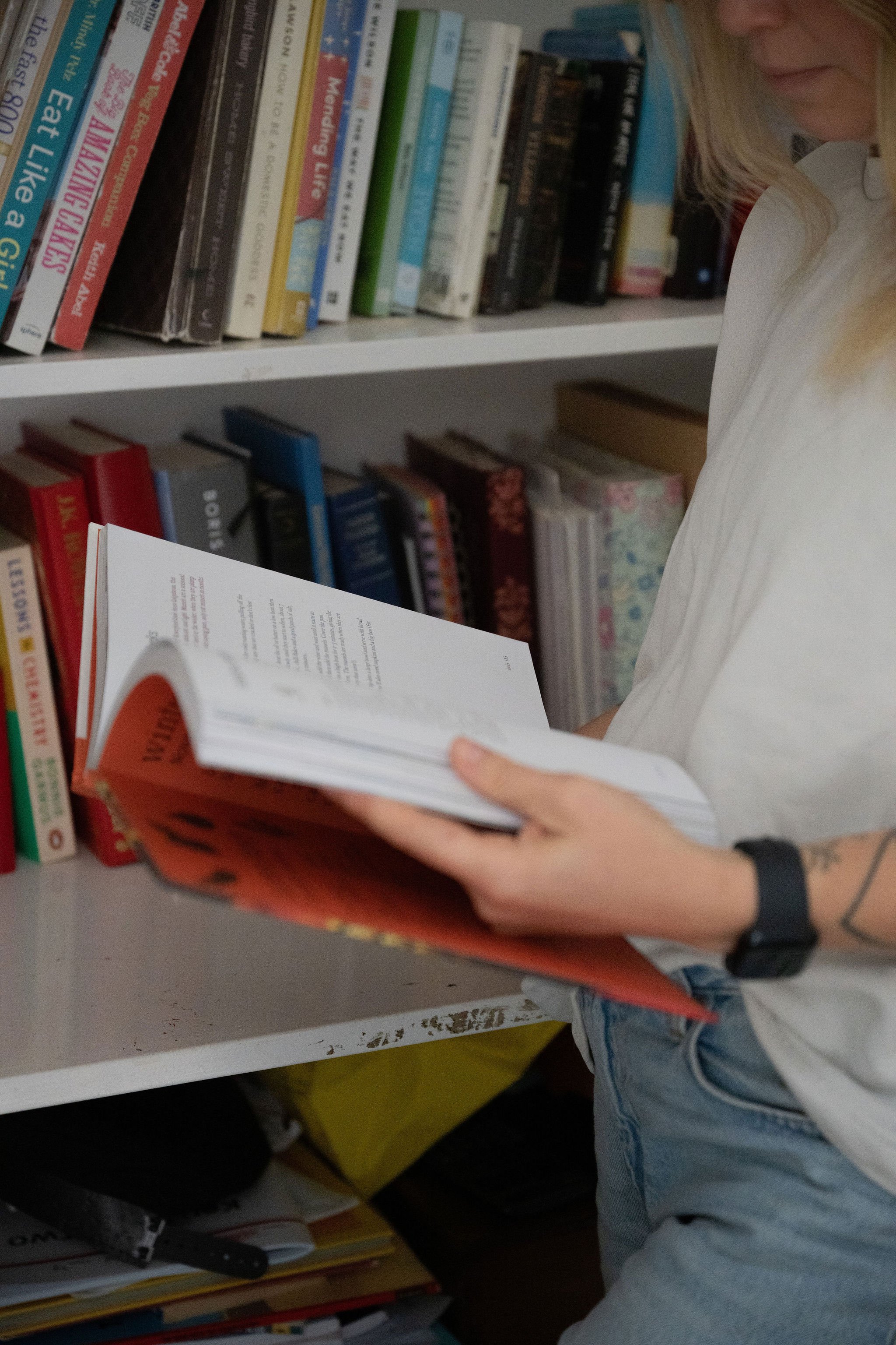 Person standing in front of a bookshelf reading a book, wearing a white shirt, jeans, and a black watch.