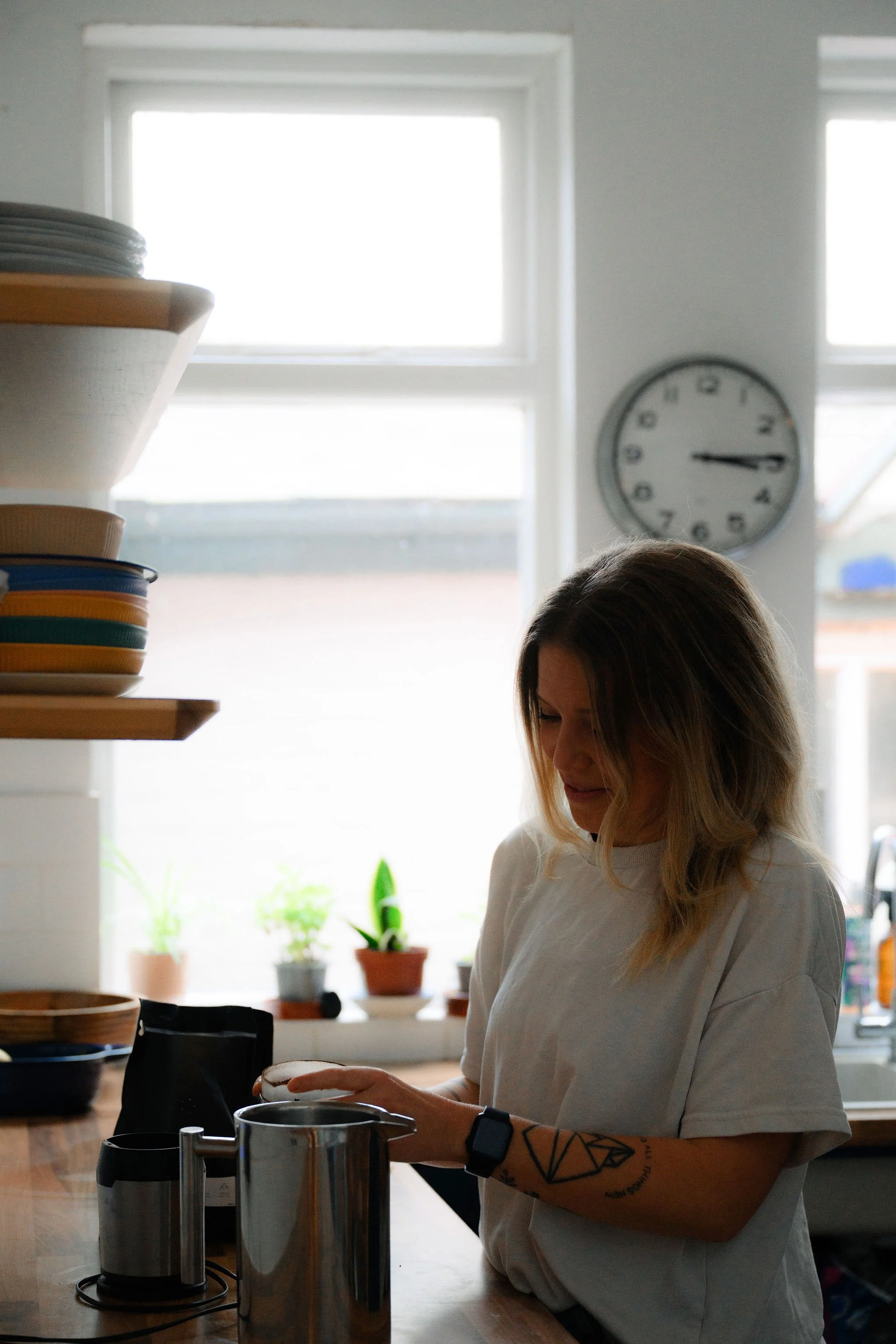 A woman with blond hair and a tattoo on her arm is preparing coffee or tea in a kitchen, standing in front of a counter with various kitchen items. There are potted plants on the window sill behind her, and a clock on the wall shows approximately 3:15.