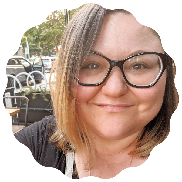 Close-up of a woman with shoulder-length brown hair, wearing glasses, smiling, sitting outdoors at a cafe or restaurant.