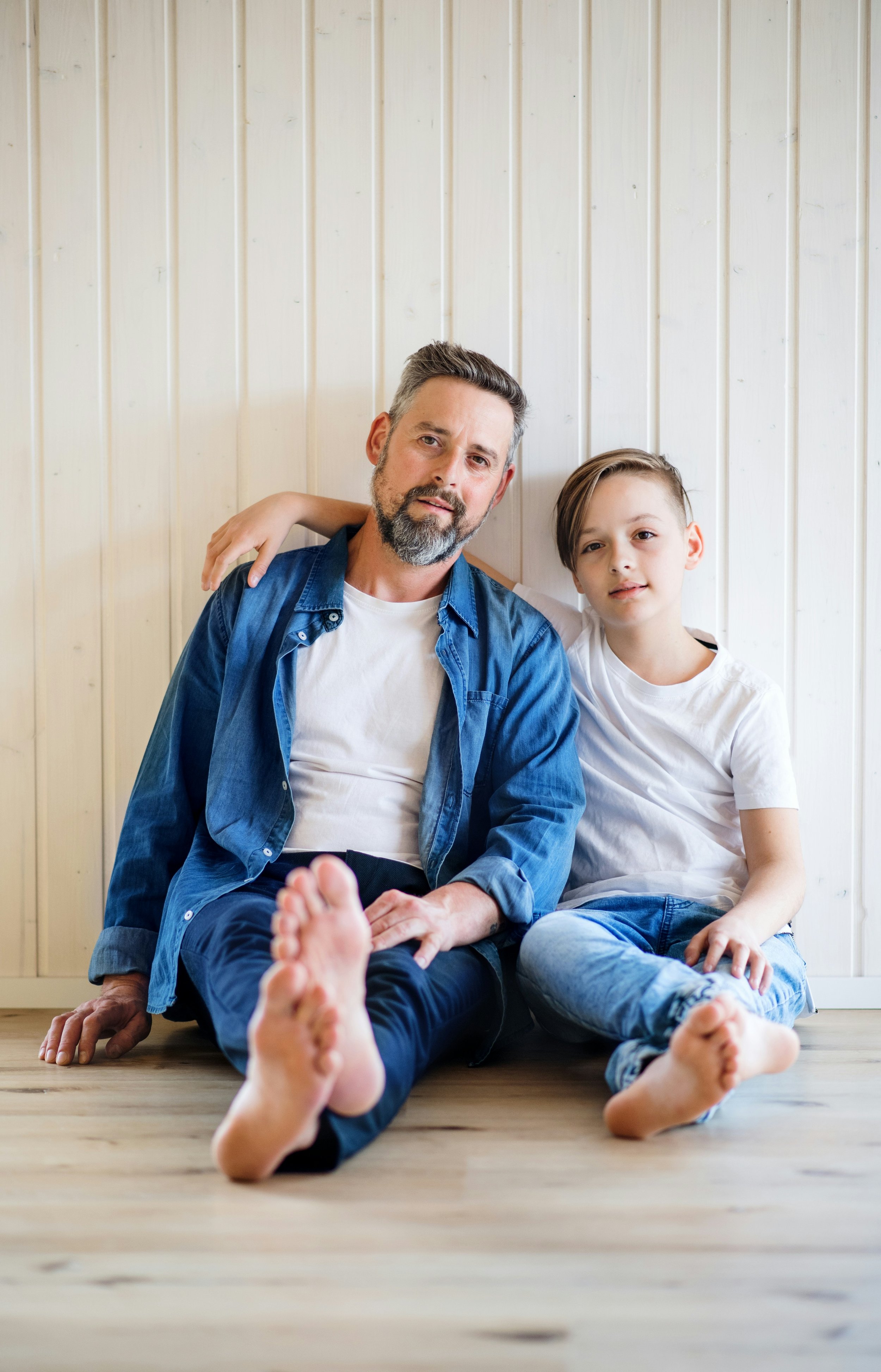A man and a boy sit on a wooden floor against a white paneled wall, with the boy's arm around the man's shoulder, both wearing casual white t-shirts and jeans.