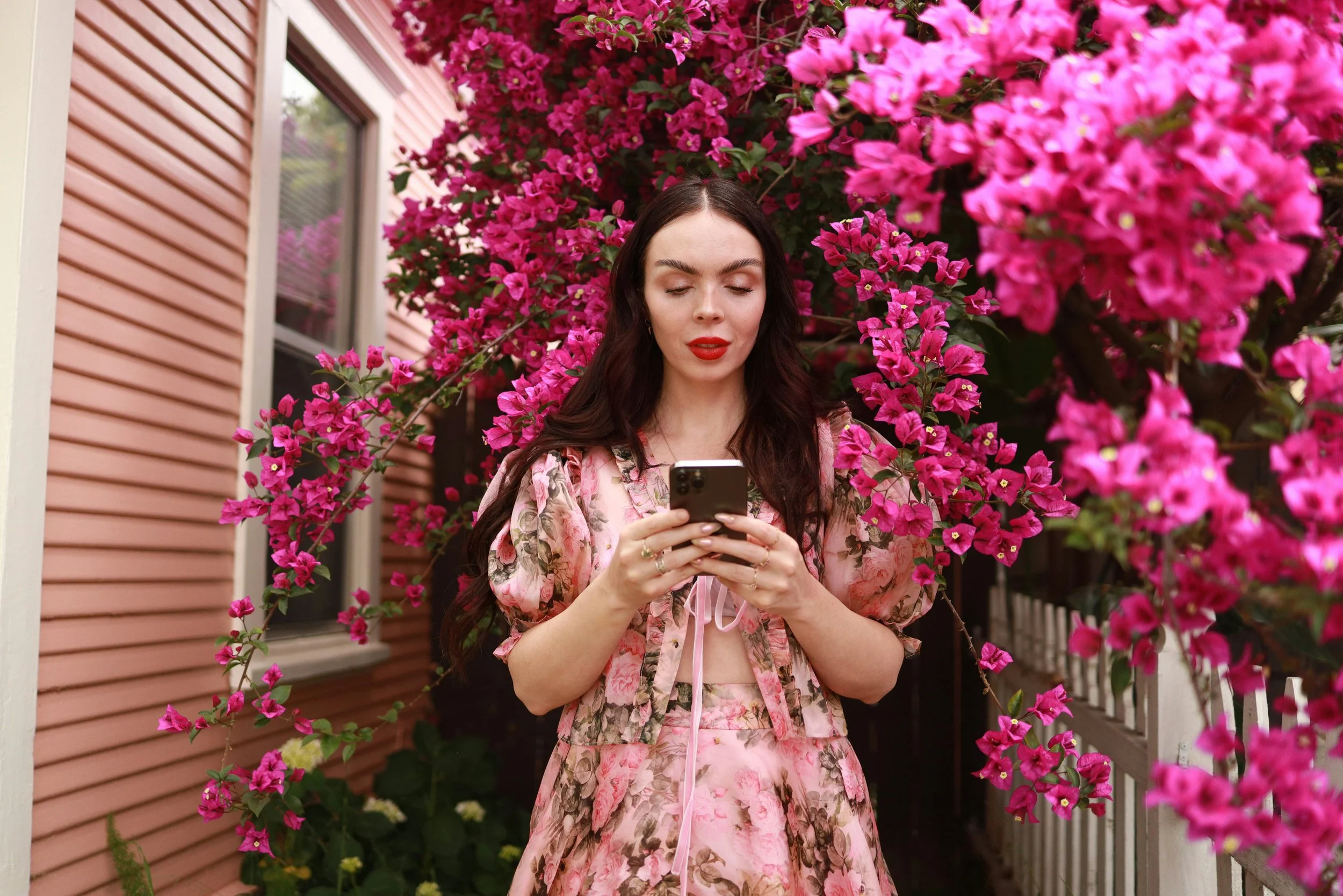 A neurodivergent woman holding a phone. She's wearing a pink dress and surrounded by bright pink bougainvillea flowers.