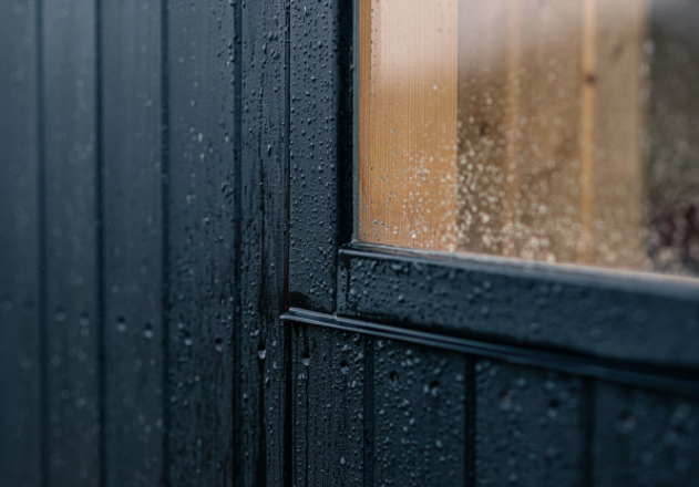 Close-up of a rainy window with water droplets on glass, dark wooden wall outside, and a faint view of a sunny outdoor scene.