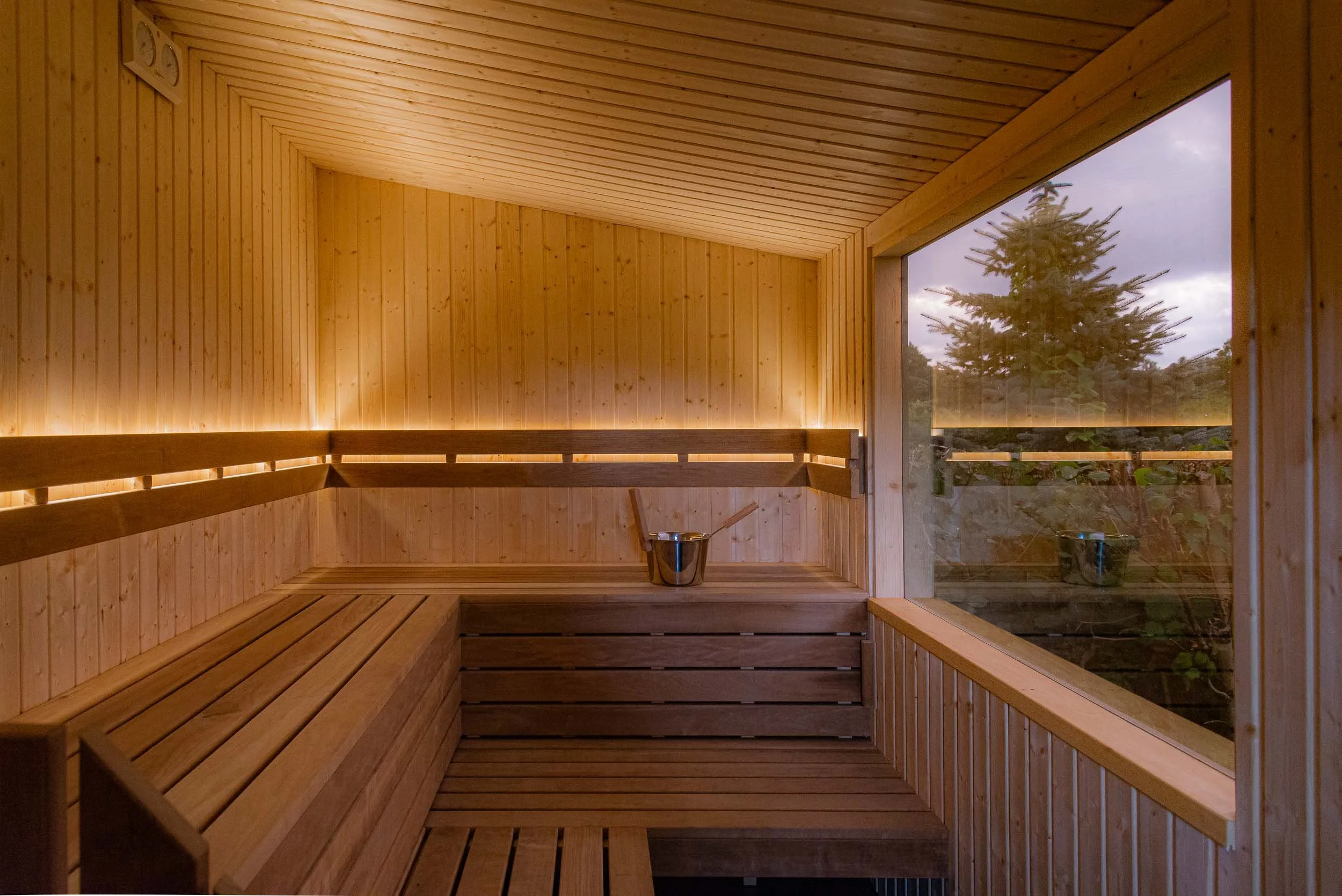 Wood-paneled sauna with a large window, benches, and a bucket with a ladle inside.