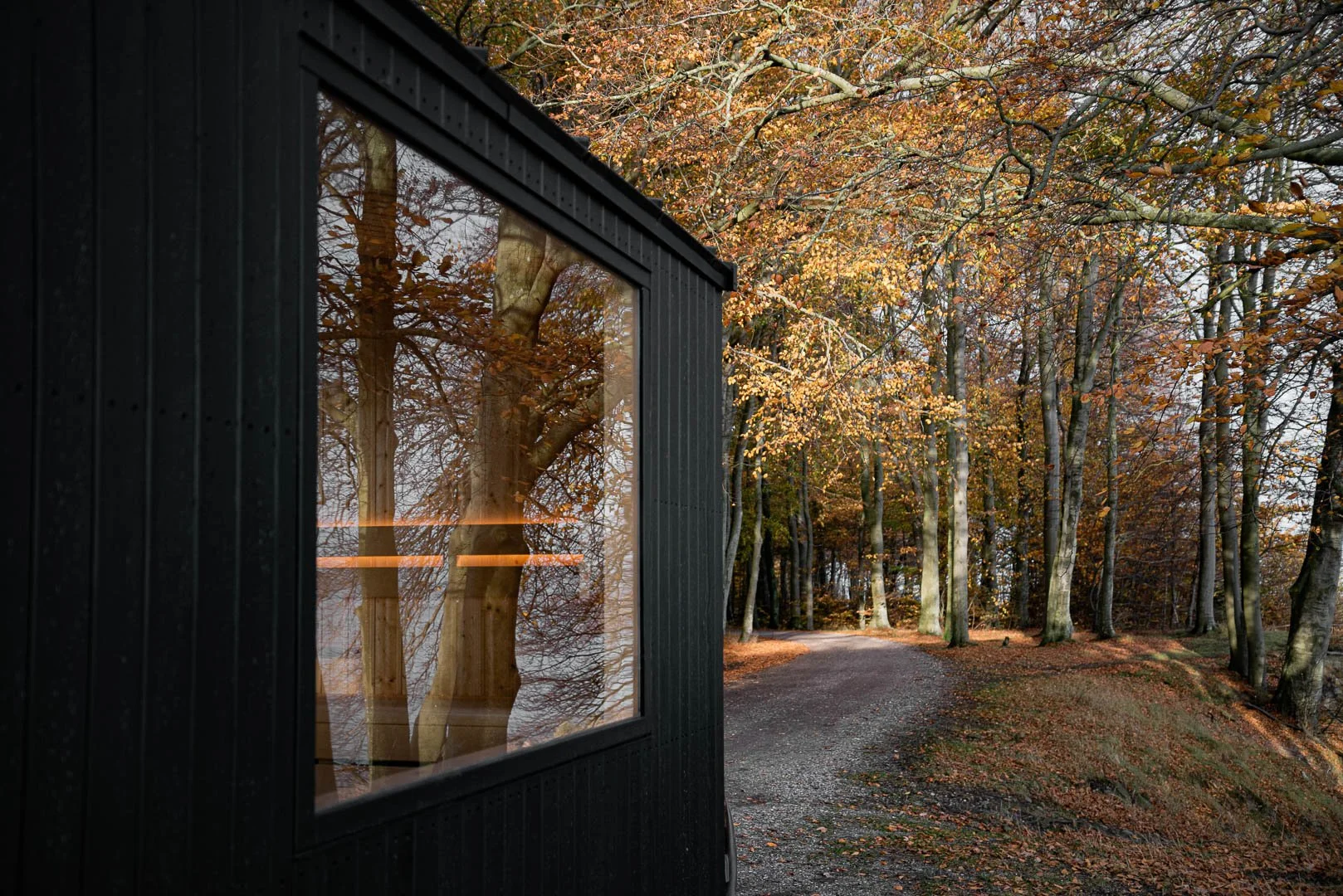 A small black cabin with a large window reflecting the autumn trees and leaves outside, situated along a gravel path in a wooded area during fall.