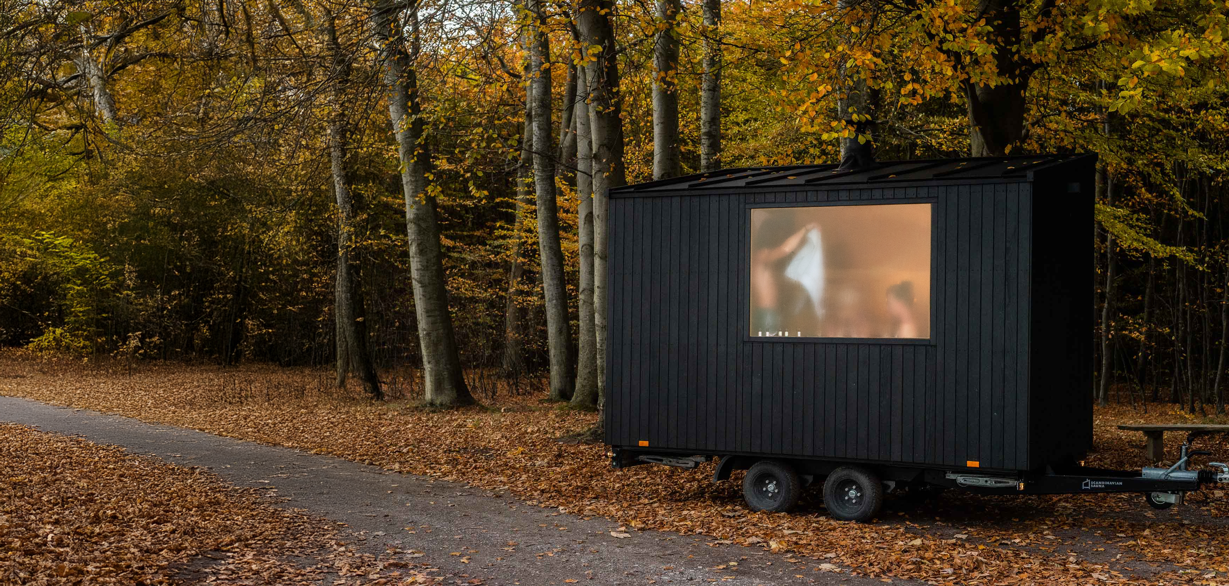 A small black trailer with a large frosted window, parked on a dirt path covered with fallen leaves, surrounded by autumn trees.