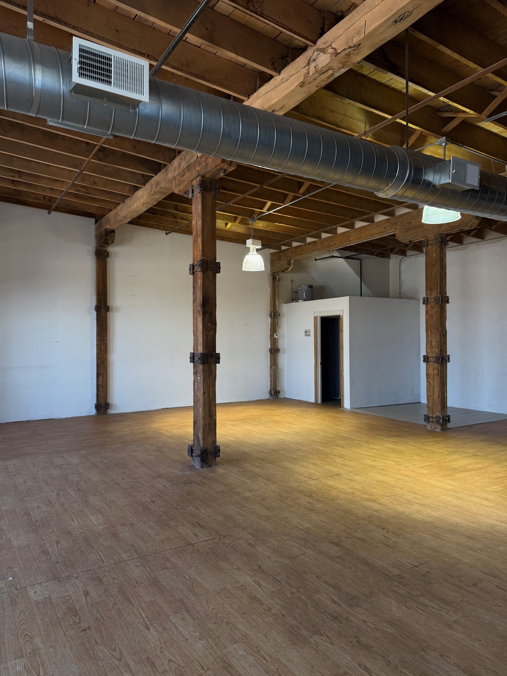 Empty industrial-style room with wooden beams, a large duct, and exposed pipes.