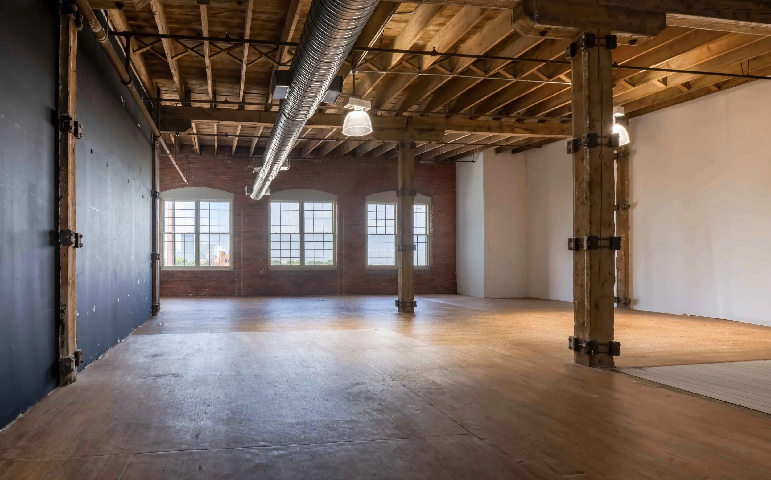 Empty industrial-style loft with wooden flooring, exposed brick wall, large multiple-pane windows, and visible wooden ceiling beams and support columns.