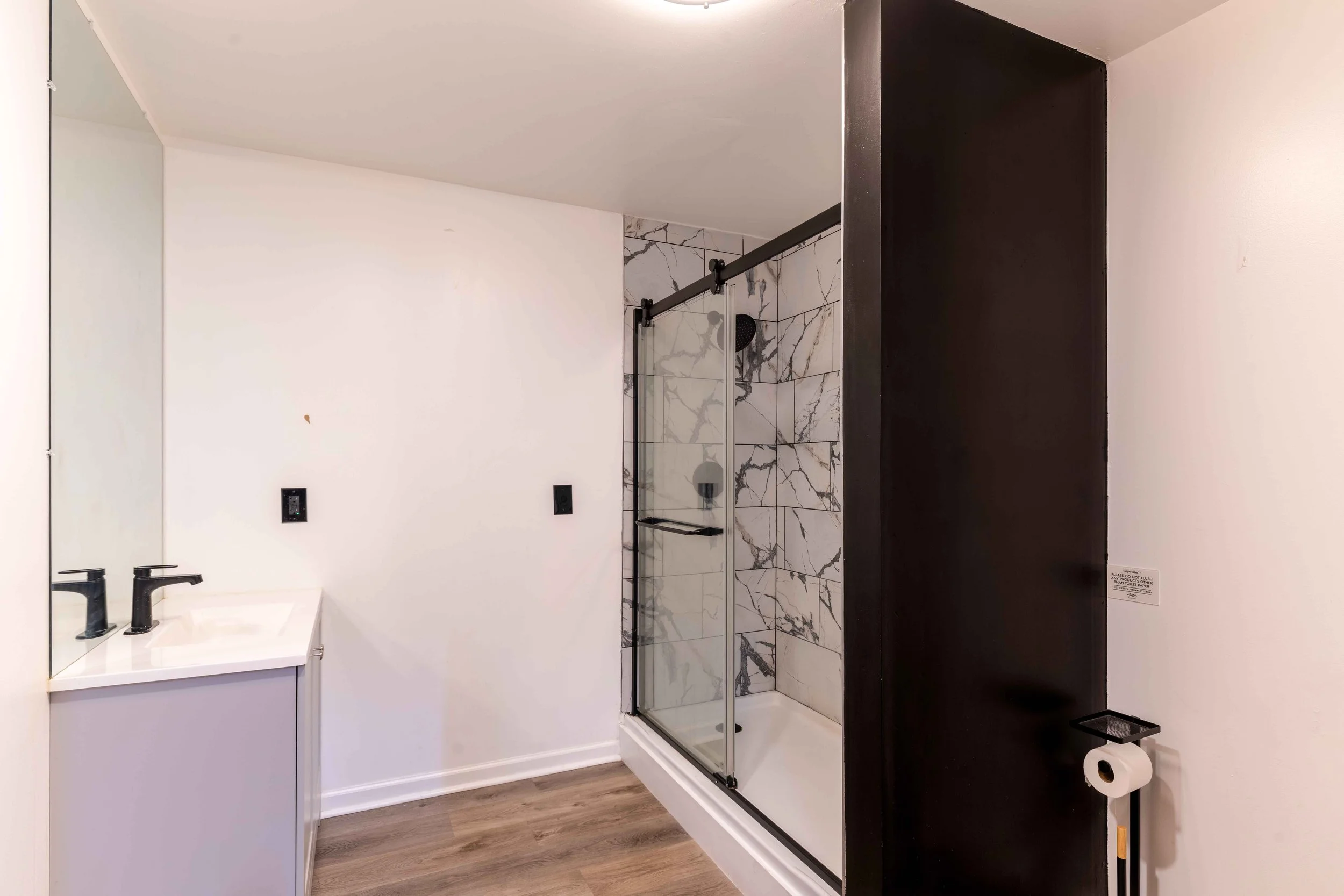 Modern bathroom featuring a white vanity with black fixtures, a framed mirror, and a glass shower with marble tiles, a black showerhead, and black sliding door hardware. The bathroom has wood-style flooring and a partially visible toilet paper holder