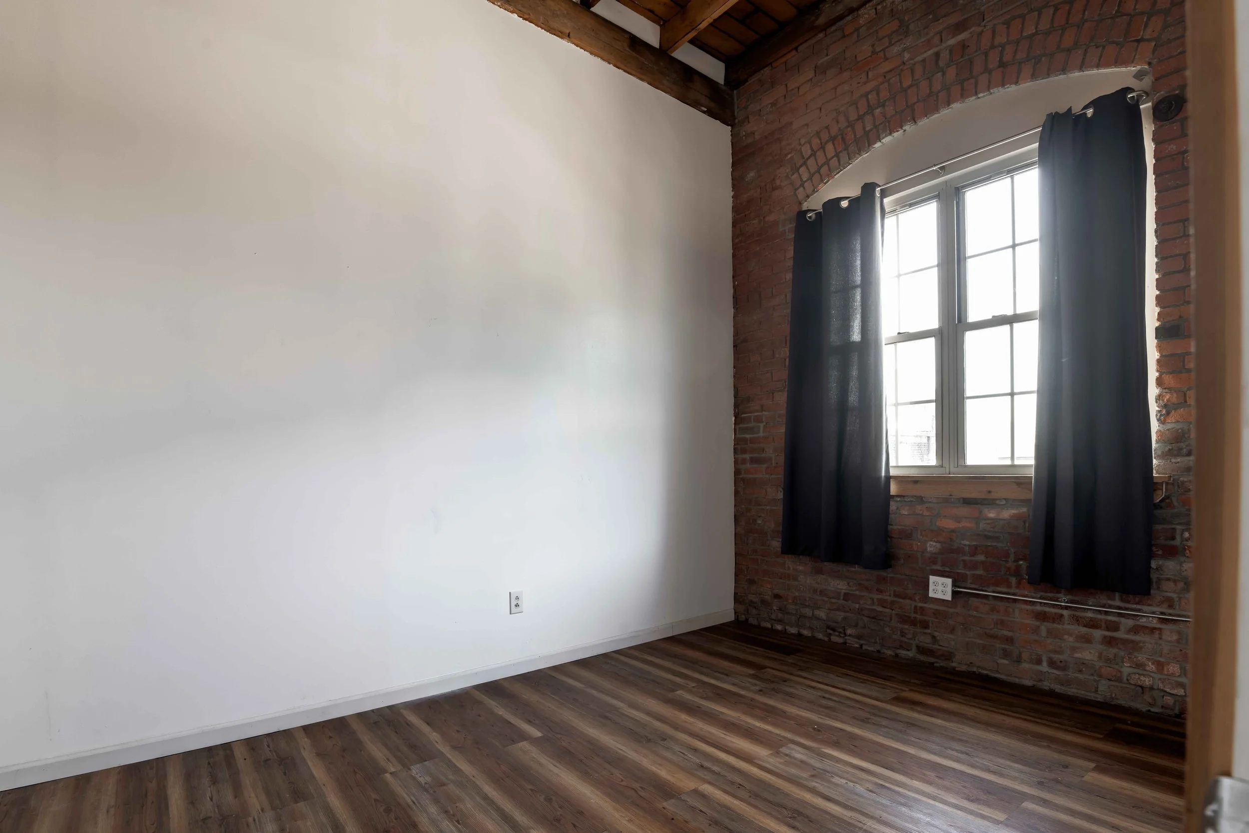 An empty room with a white wall and a brick wall with a window with black curtains, hardwood flooring, and exposed wooden beams on the ceiling.