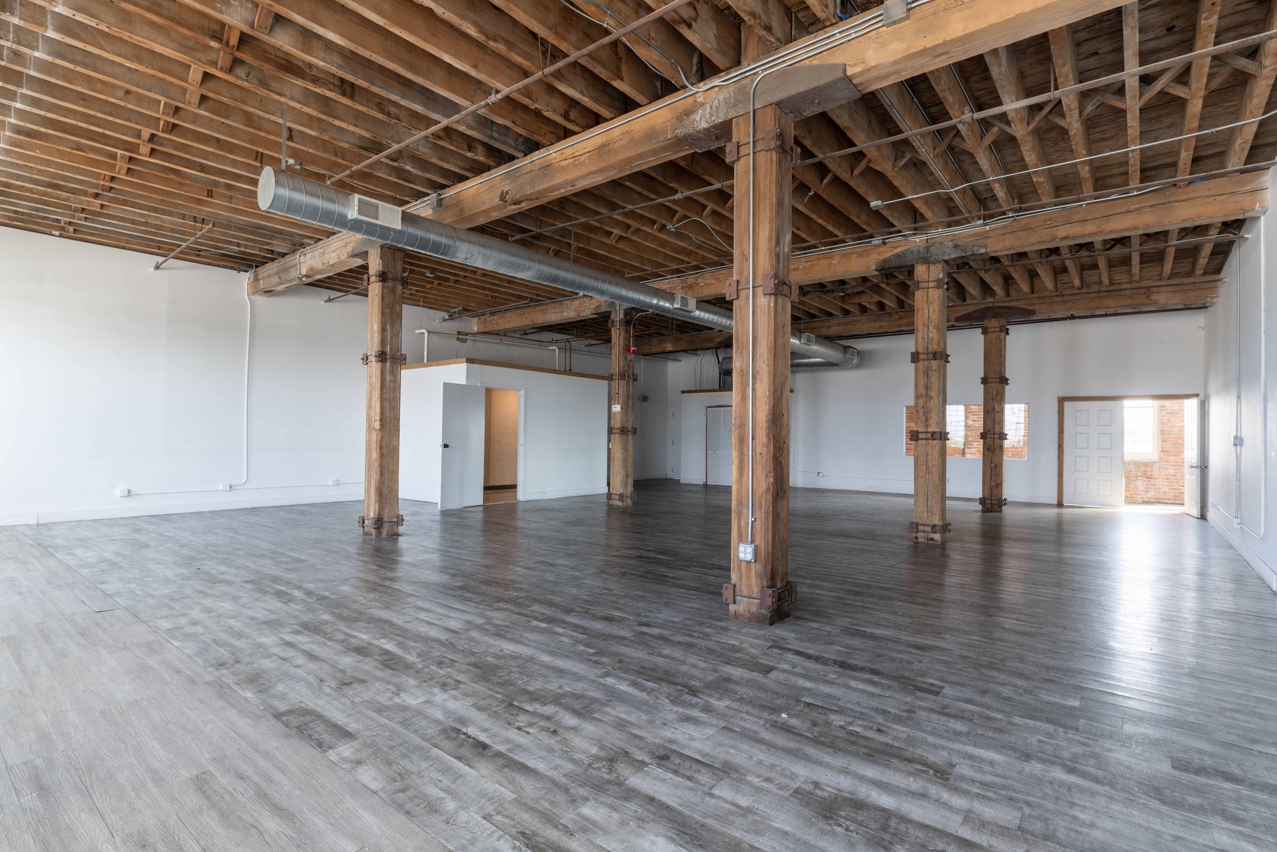 Empty interior with exposed wooden ceiling beams, white walls, and wooden flooring, featuring structural wooden columns and several doors and windows.