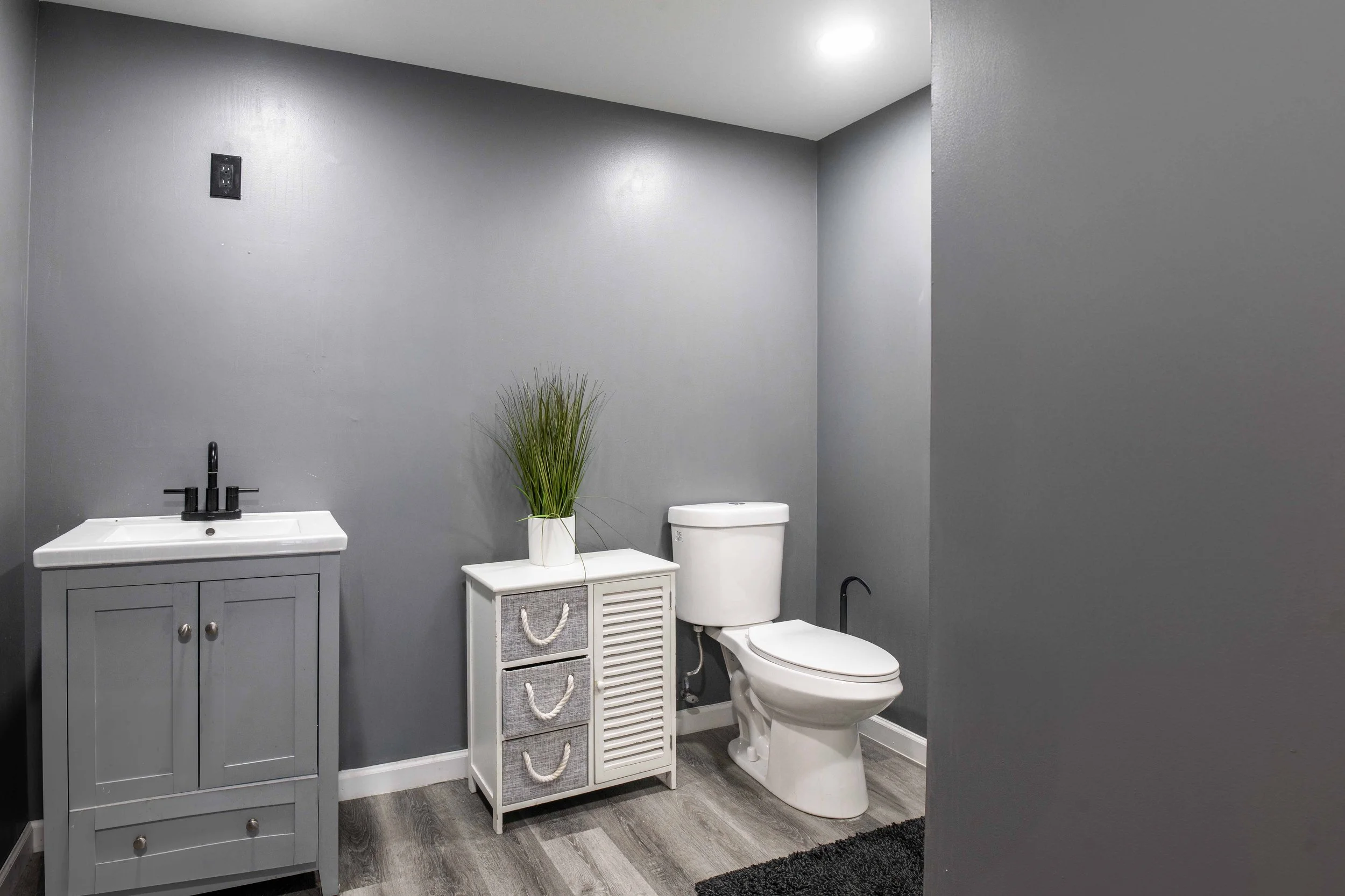 A small bathroom with gray walls, a white toilet, a light gray cabinet sink, a small white cabinet with three drawers and a potted plant on top, and a black rug on the floor.