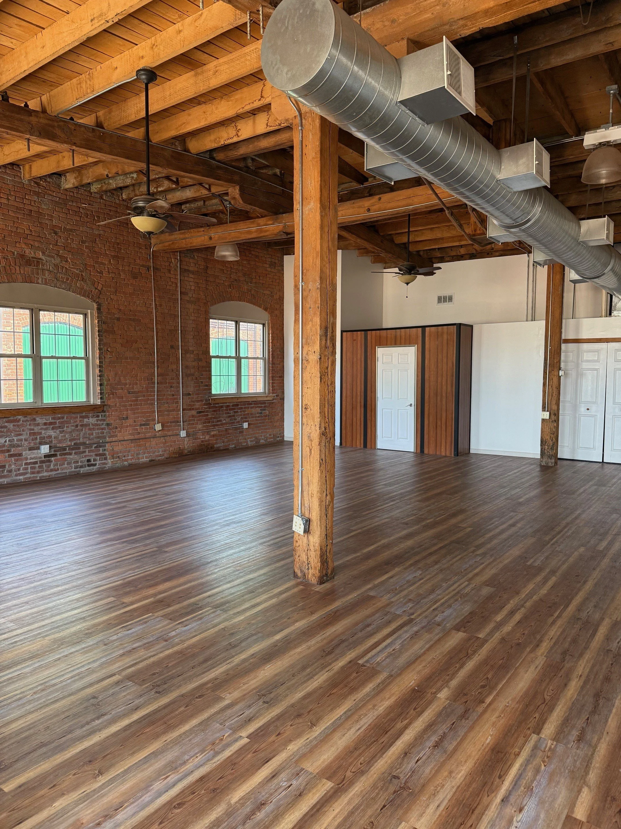 An empty industrial-style loft with wooden beams, ductwork, ceiling fans, brick walls, and wood flooring.