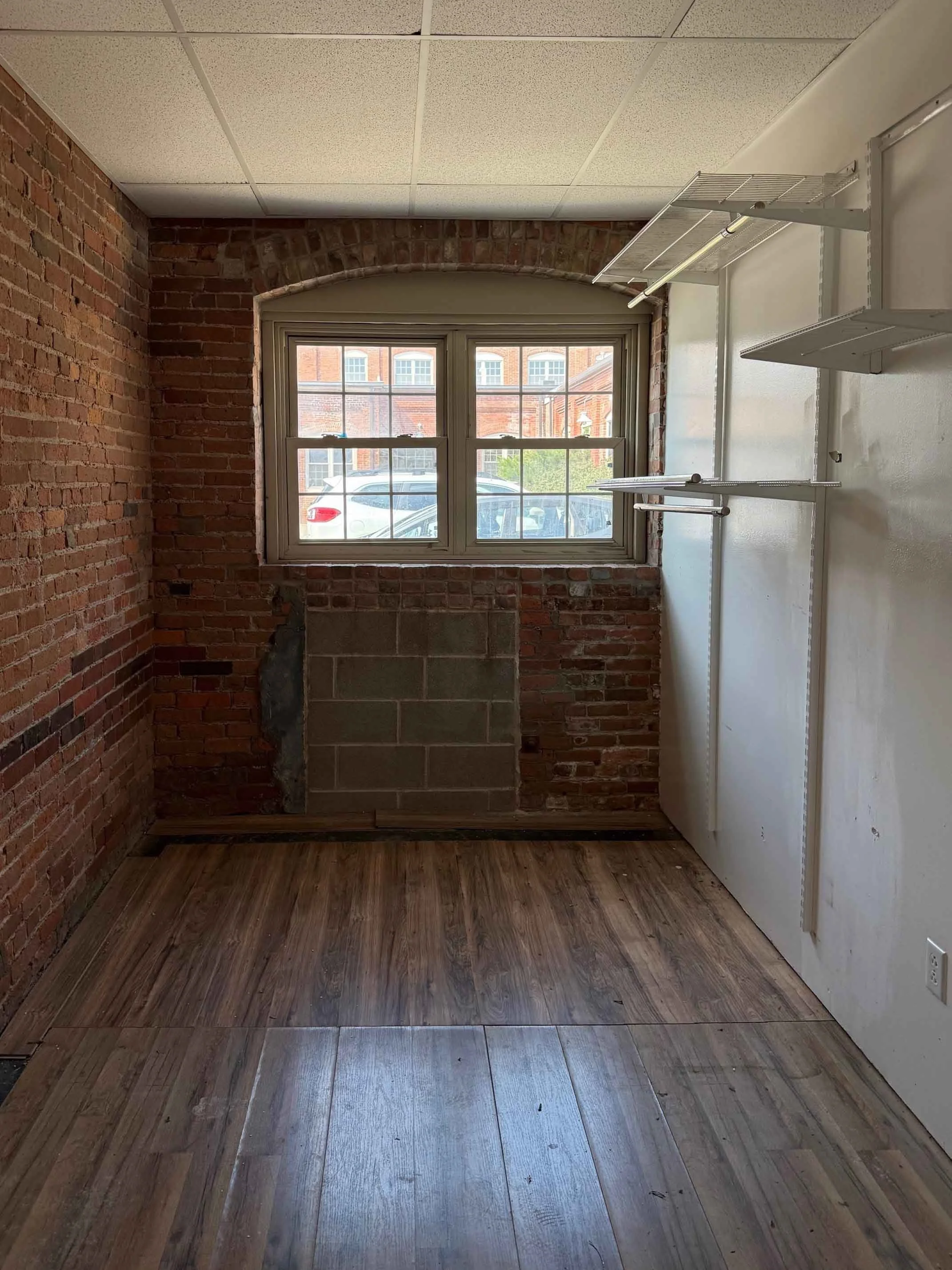 A small room with brick walls and a window at the back. Shelves are installed on the white wall to the right. The floor is partially covered with wooden planks, and there is an unfinished section on the brick wall below the window.