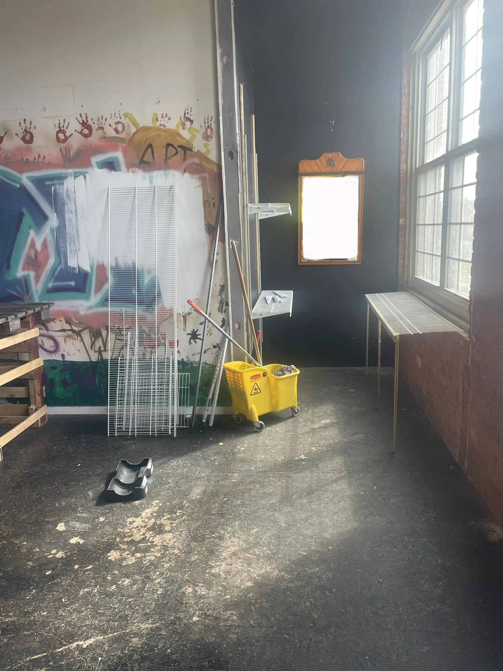 An art studio with graffiti on the wall, cleaning supplies including a mop and yellow bucket, metal racks, a wooden table, and large windows allowing natural light.