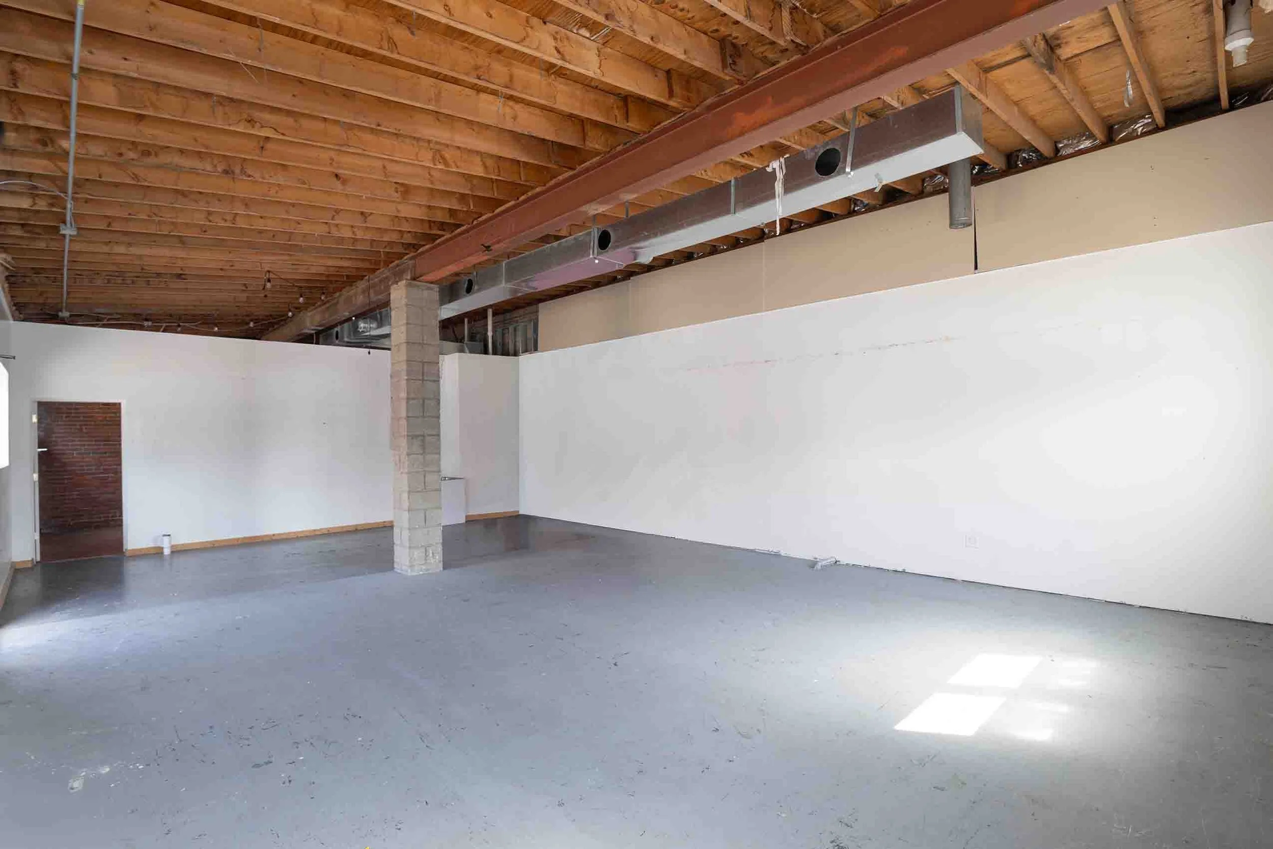 An empty industrial-style room with concrete flooring, white walls, an exposed wooden ceiling, and a metal beam. There is a small door on the left wall, and a brick column in the middle supporting the ceiling.