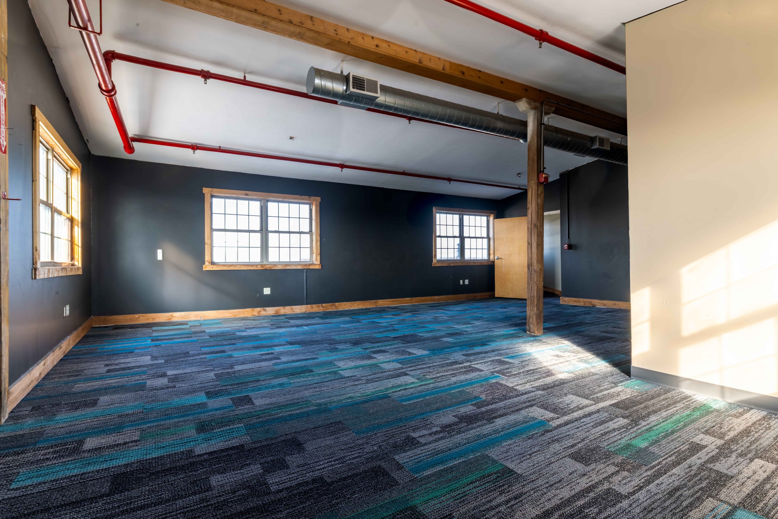 Empty office space with dark walls, wooden trim, multiple windows, carpeted floor, and ceiling pipes and ductwork.