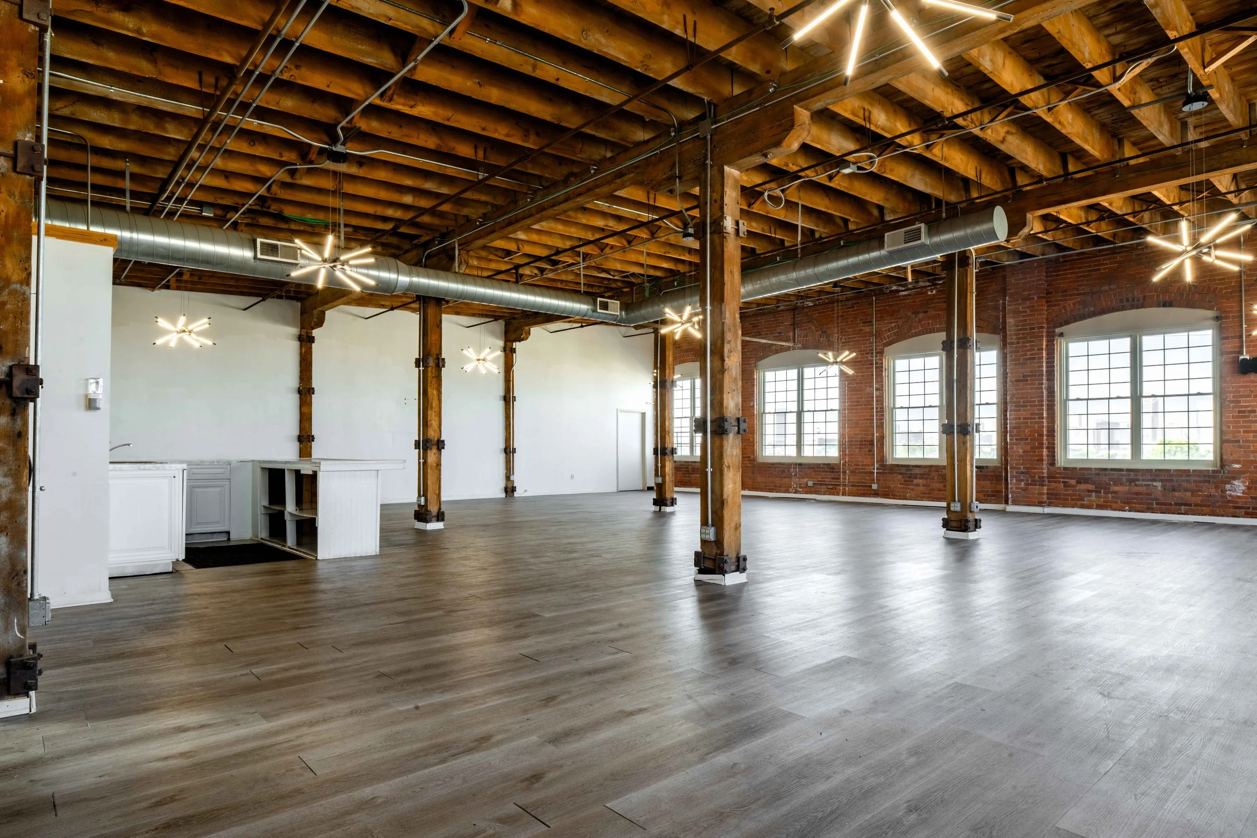 An empty industrial-style room with wooden beams, large windows, exposed brick walls, modern lighting fixtures, and a wooden floor.