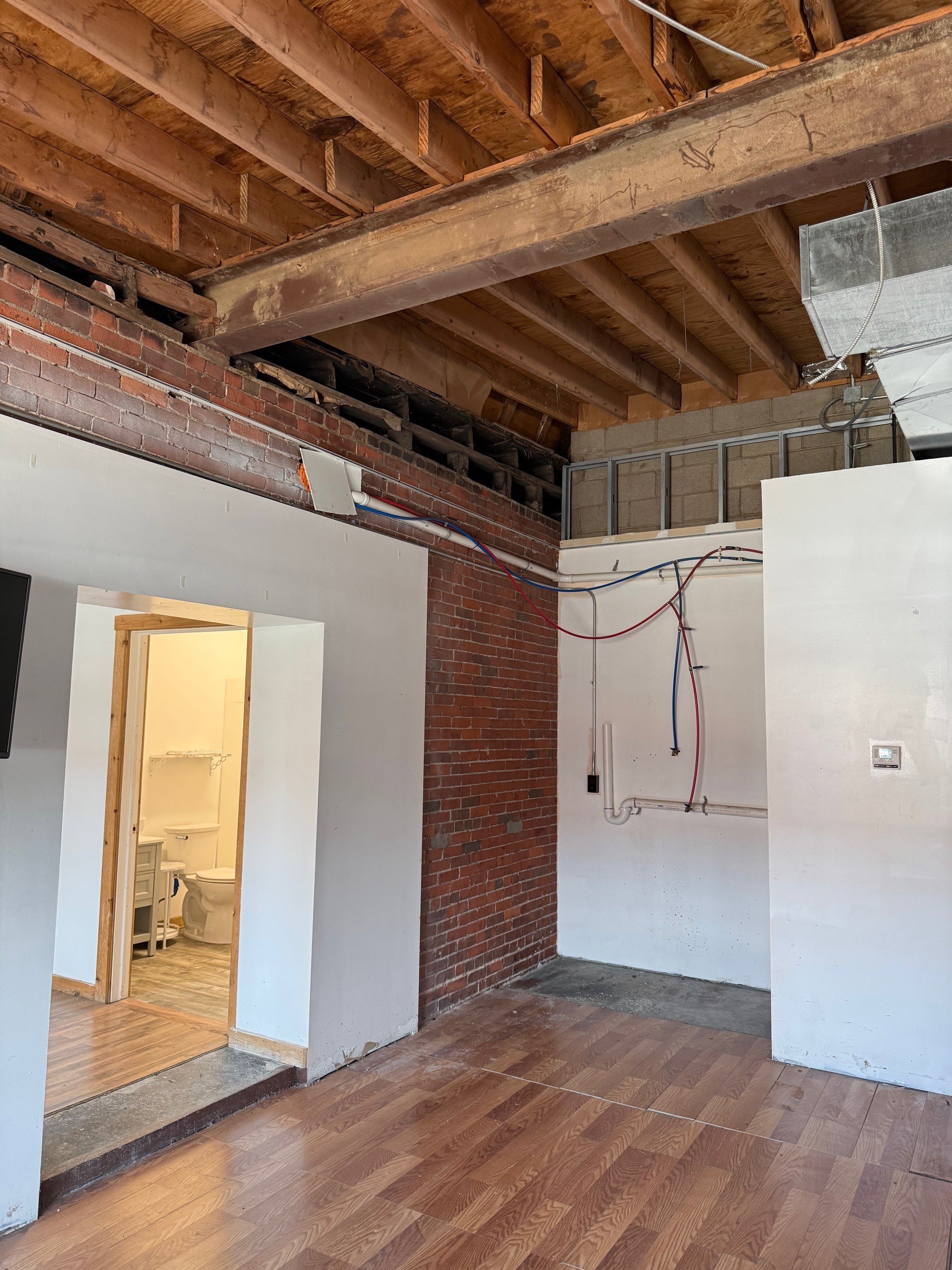 Interior of a room under renovation with exposed brick wall, electrical wiring, and unfinished ceiling, with a nearby bathroom visible through an open doorway.