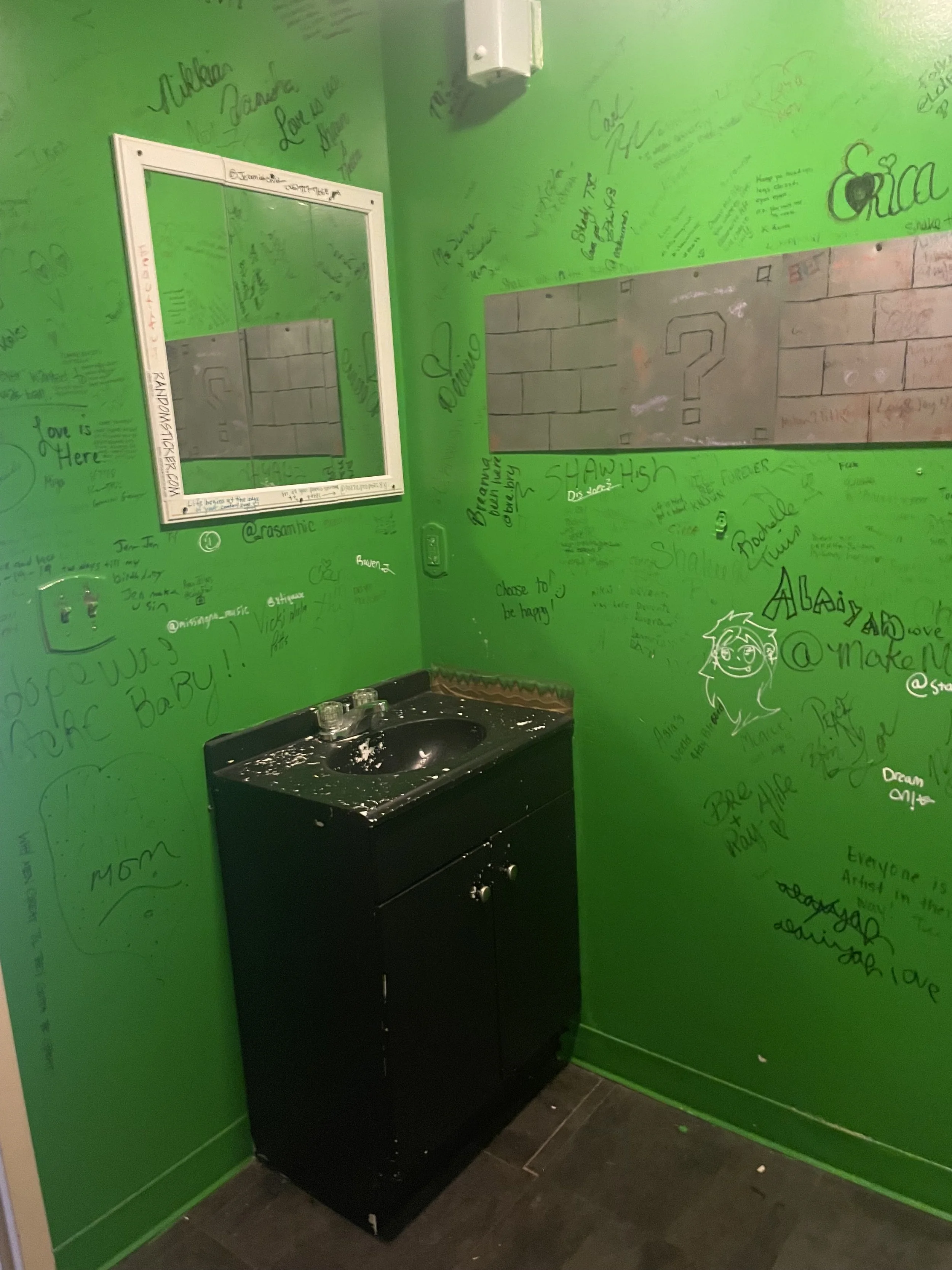 Green-painted wall with handwritten messages, signatures, and doodles, along with a mirror and a small black sink cabinet in a graffiti-covered room.