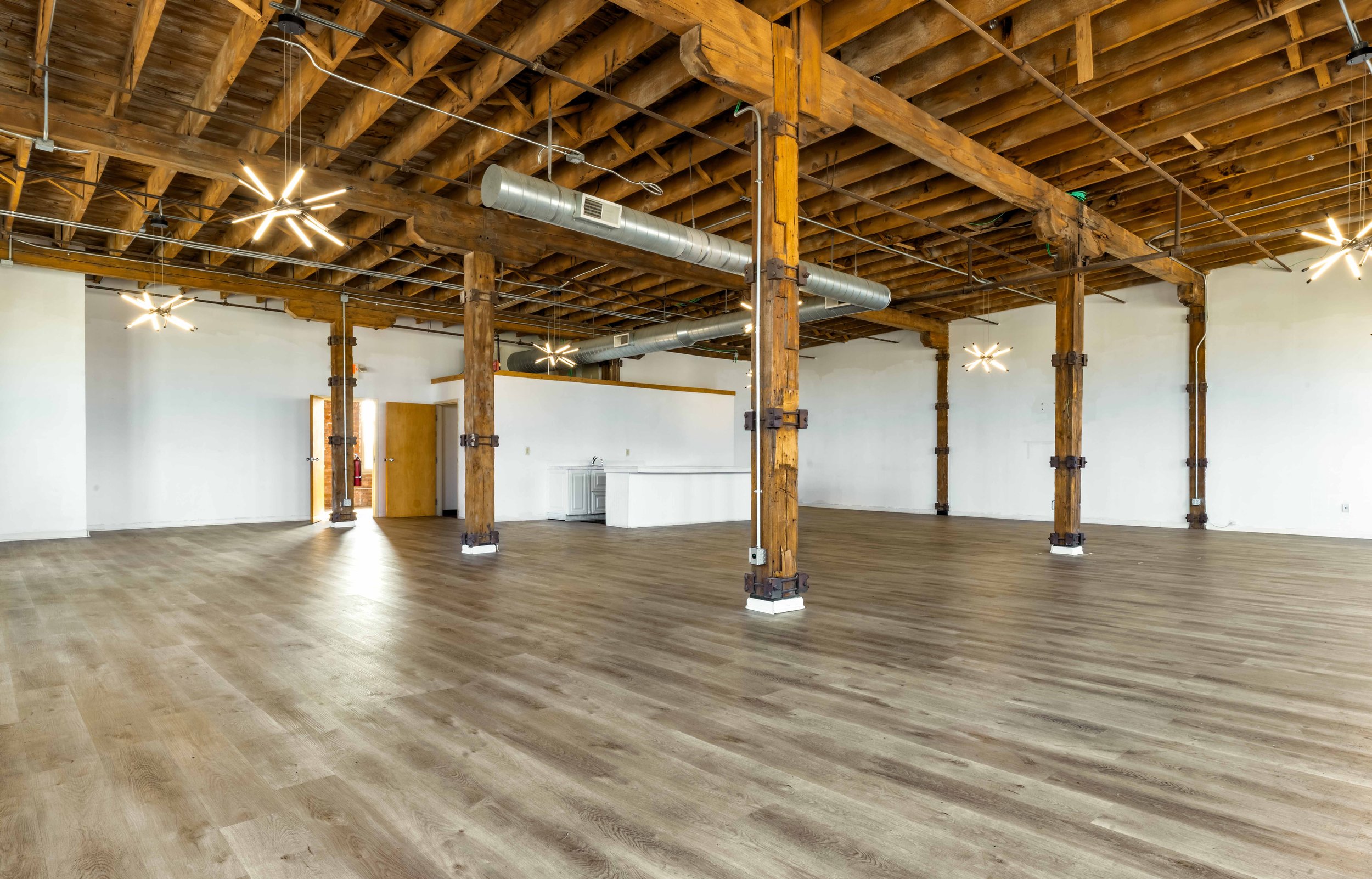 Open, spacious room with wooden floors, exposed wooden beams, and industrial-style ceiling pipes, illuminated by modern star-shaped ceiling lights, with a kitchenette area in the back and multiple empty walls.