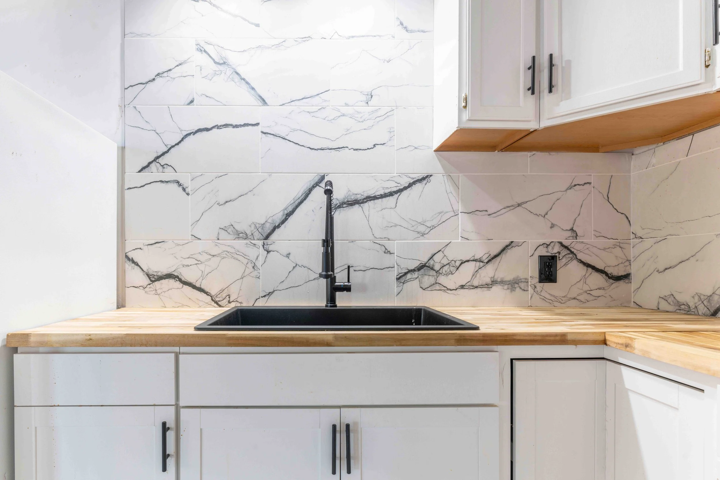 Kitchen with black sink, black faucet, marble backsplash, white cabinets, and a wood countertop.