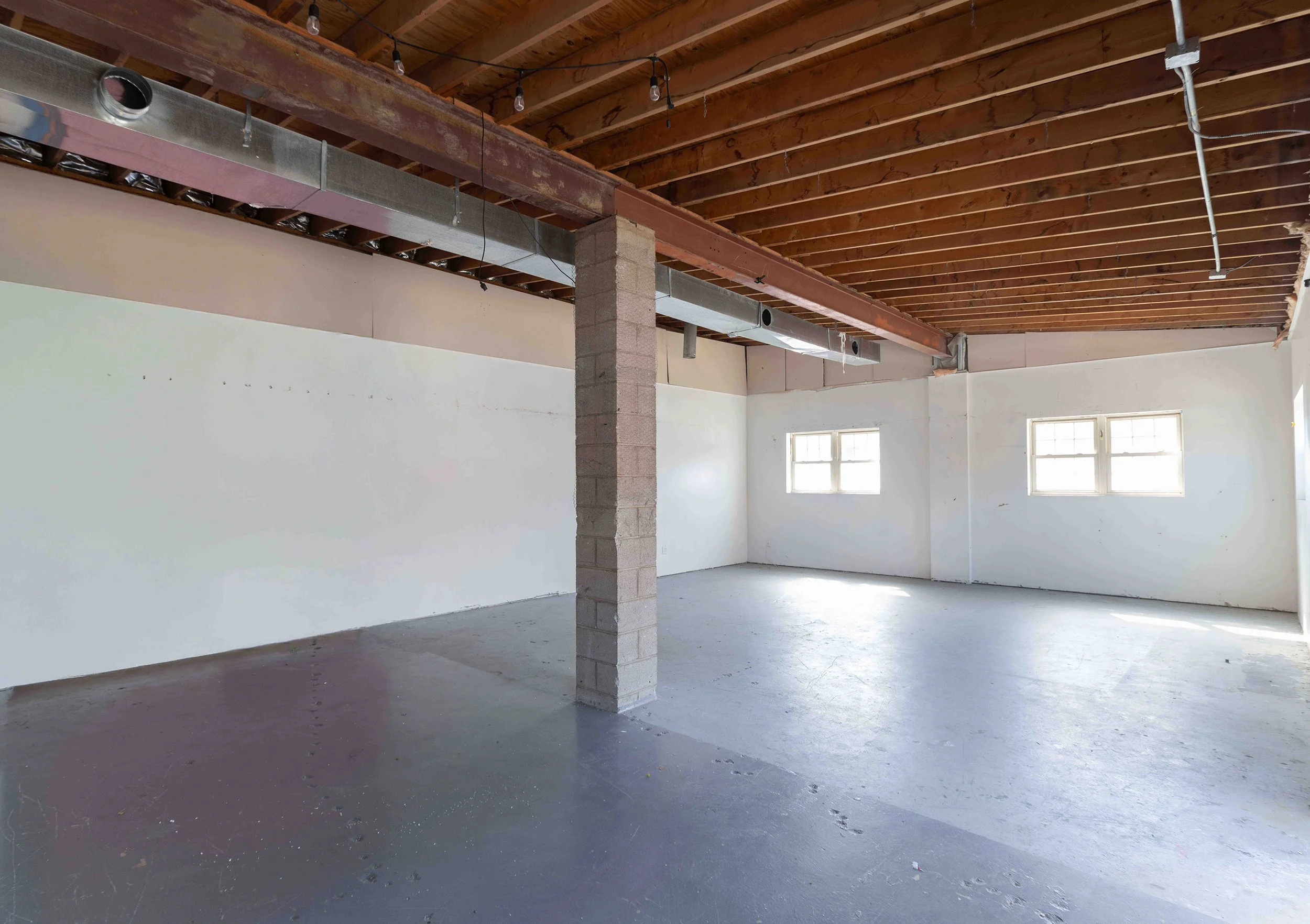 Empty interior room under construction with white walls, unfinished wooden ceiling with exposed beams, a concrete support column, and small windows letting in natural light.