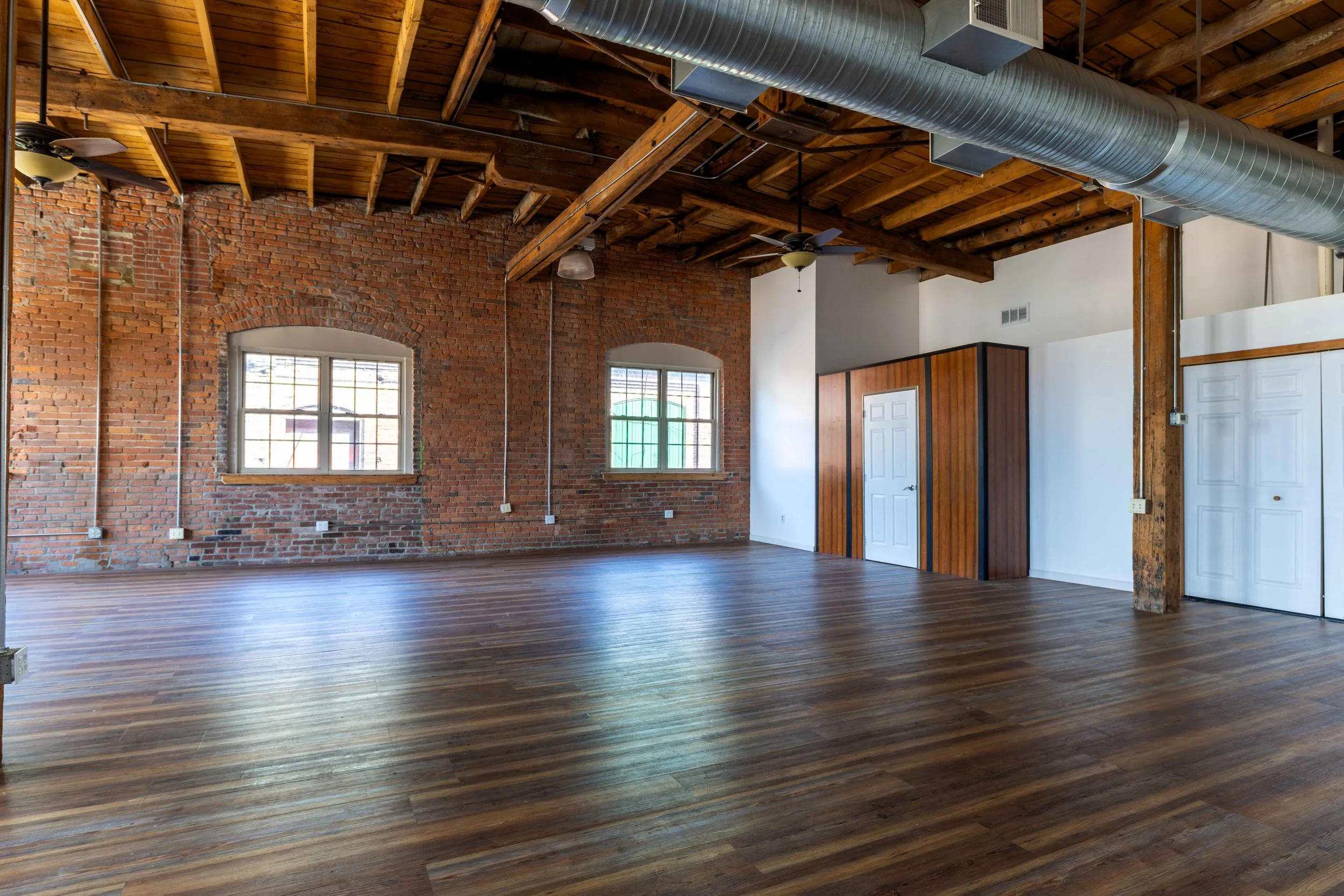 Empty loft-style room with exposed brick walls, wooden ceiling and floor, large windows, and visible ductwork.