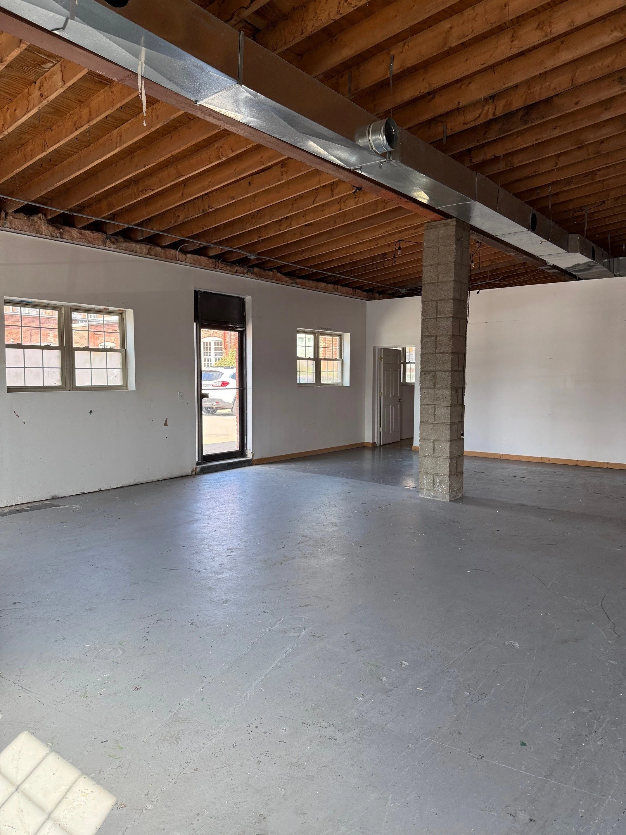 Empty storefront with large open space, gray concrete floor, white walls, exposed wooden ceiling, and three windows and a door.