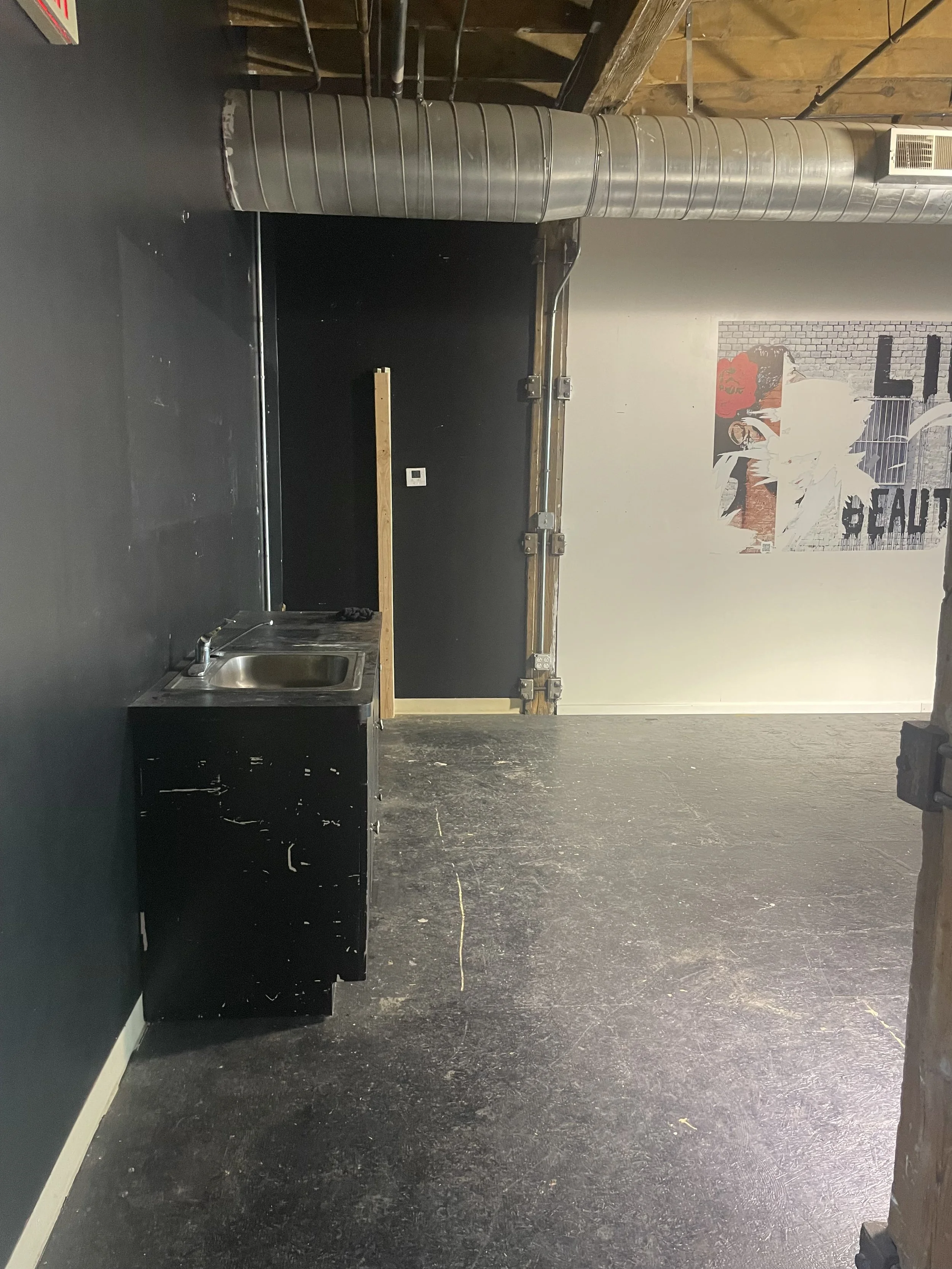 An empty room with black walls, a small stainless steel sink, a black cabinet, a wooden board, a black door, a wall poster, and exposed industrial ductwork on the ceiling.