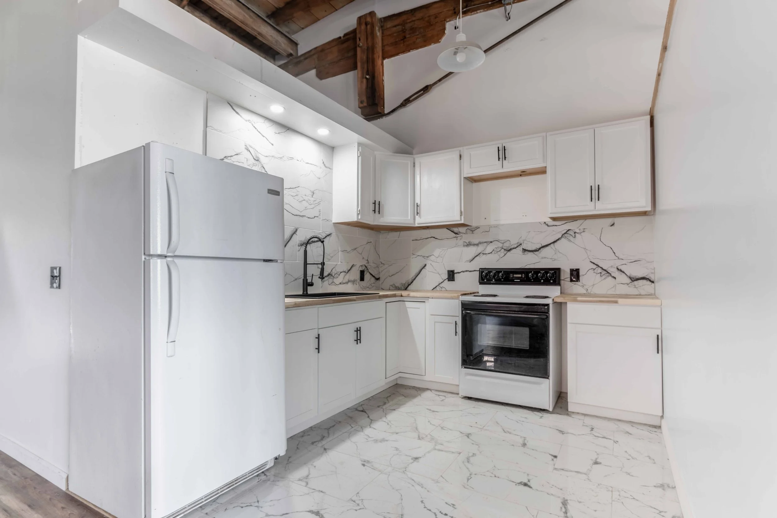 Modern kitchen with white cabinets, black hardware, black faucet, marble backsplash, marble floor, white refrigerator, and black stove, in a room with exposed ceiling beams.