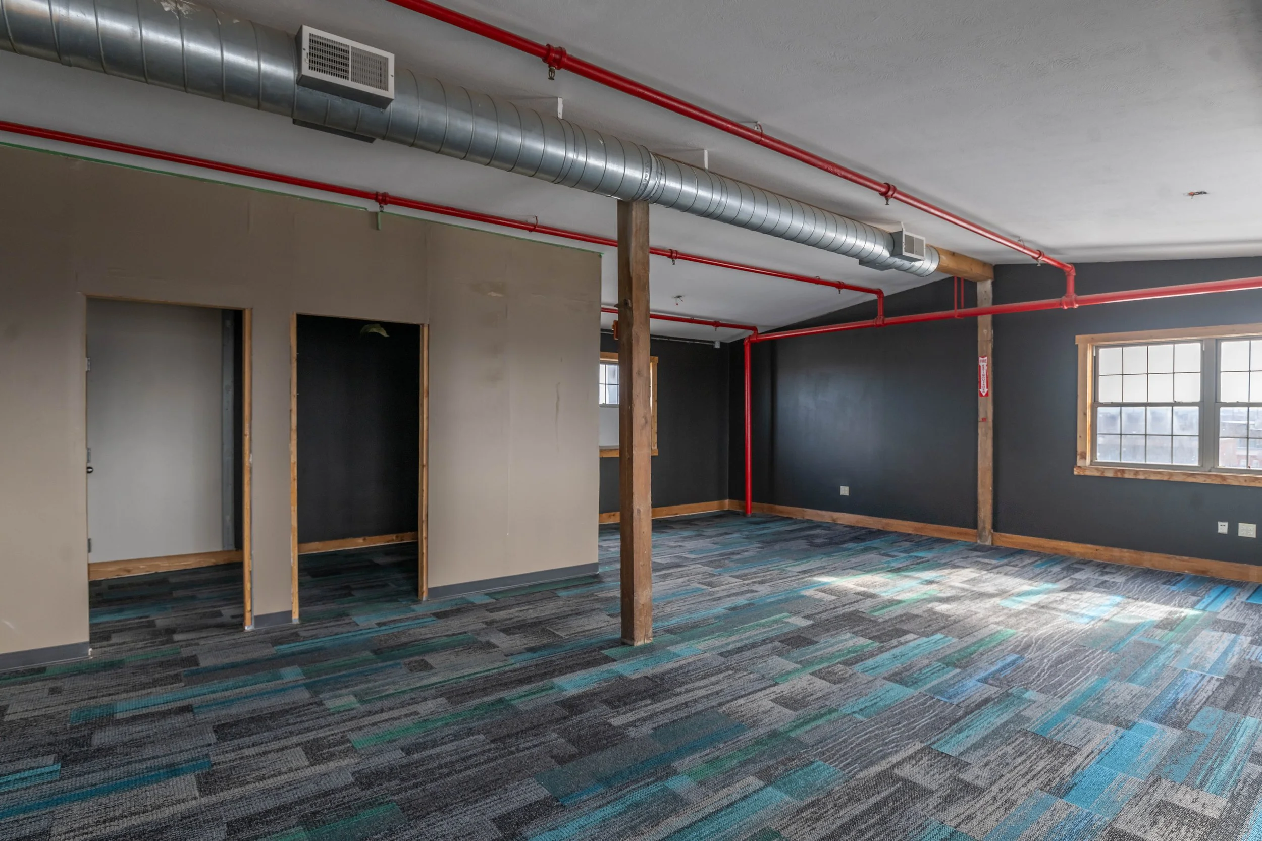 Unfinished commercial interior with exposed ductwork, wooden support beams, dark and beige walls, and windows with natural light, along with a patterned carpeted floor.