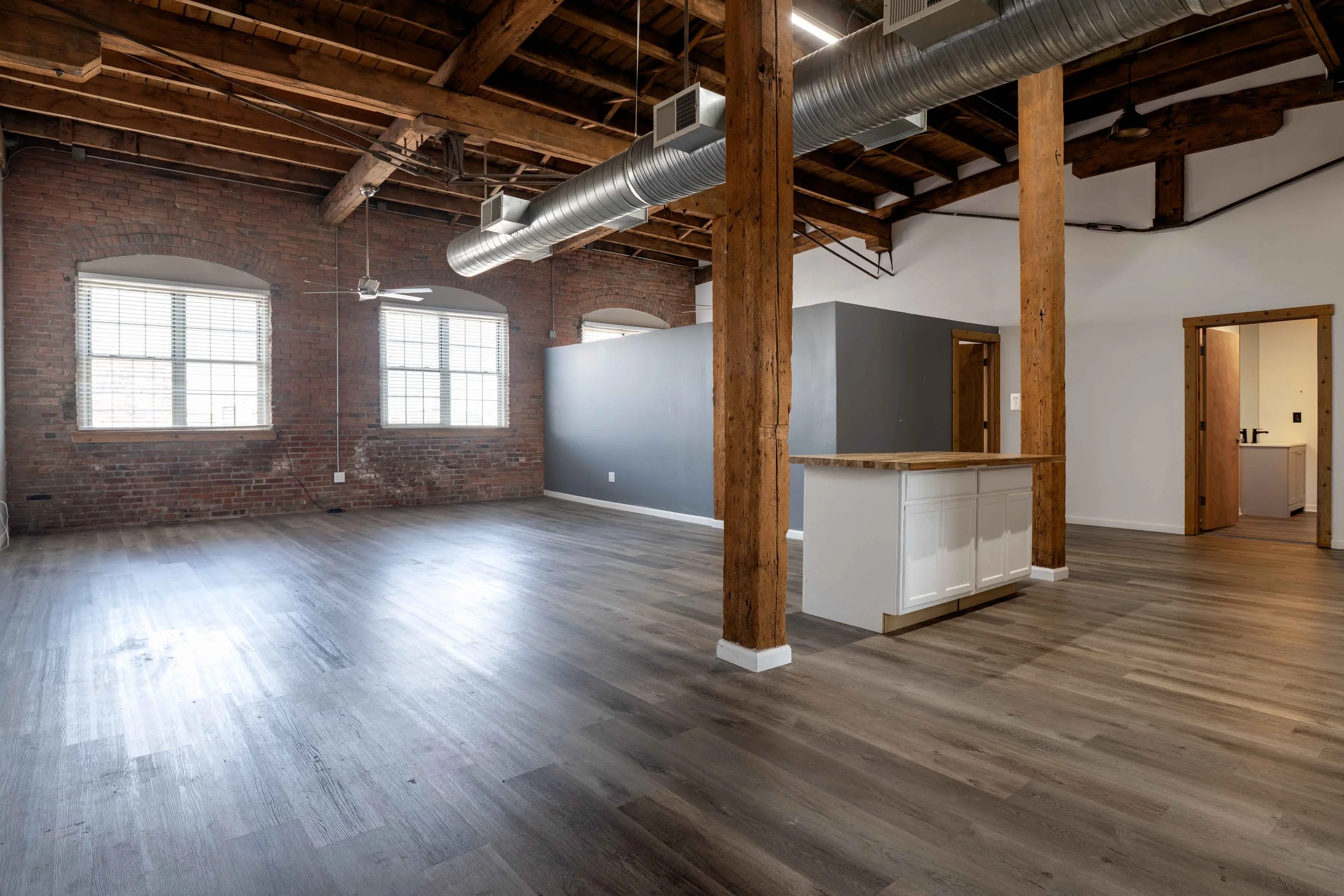 Spacious open loft with exposed brick walls, wooden beams, hardwood flooring, and large windows, featuring a small kitchen area with white cabinets and an island.