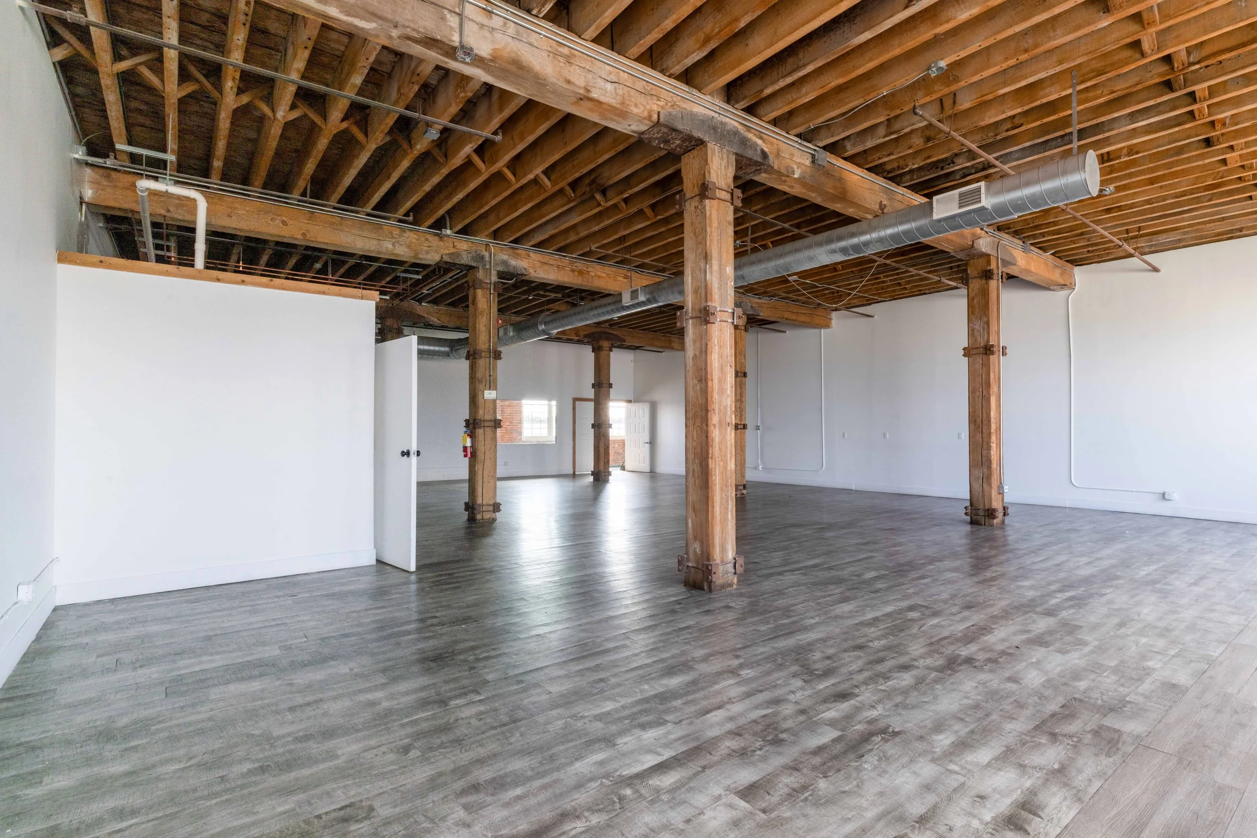 Empty interior space with exposed wooden beams and columns, wood flooring, white walls, and a ventilation duct running across the ceiling.