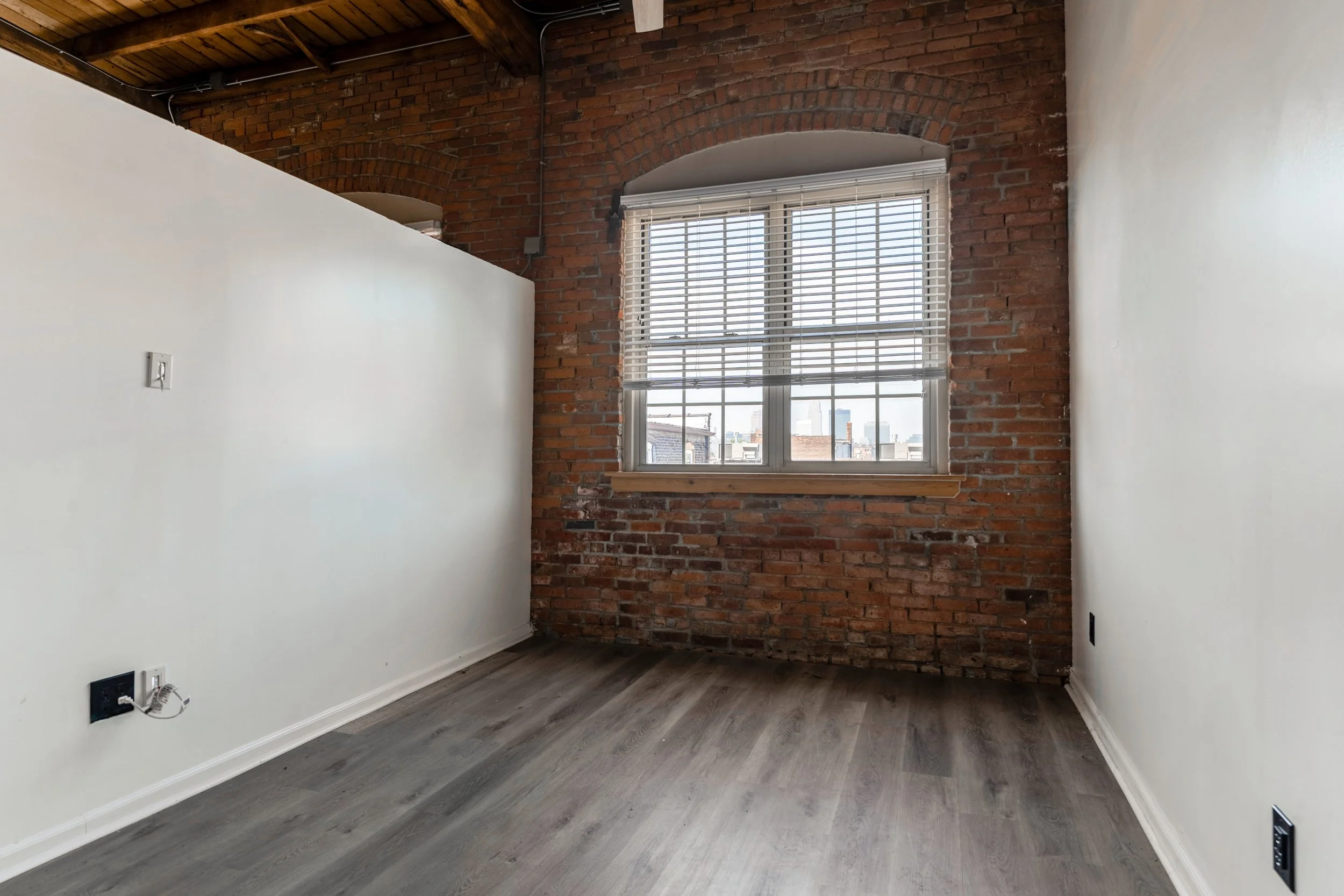 Empty room with white walls, a large window with blinds, brick accent wall, and wood flooring.