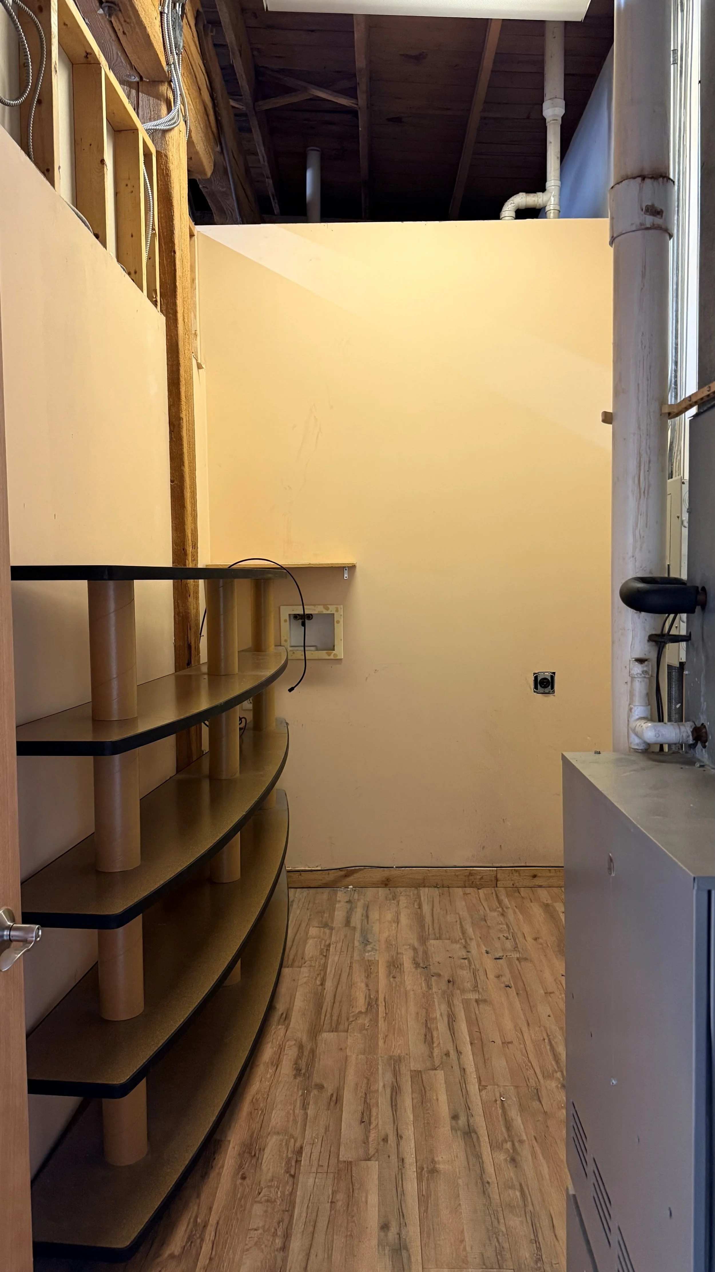 Construction area with partially finished room, open wooden beams ceiling, beige wall, wooden floor, and shelves on left side.