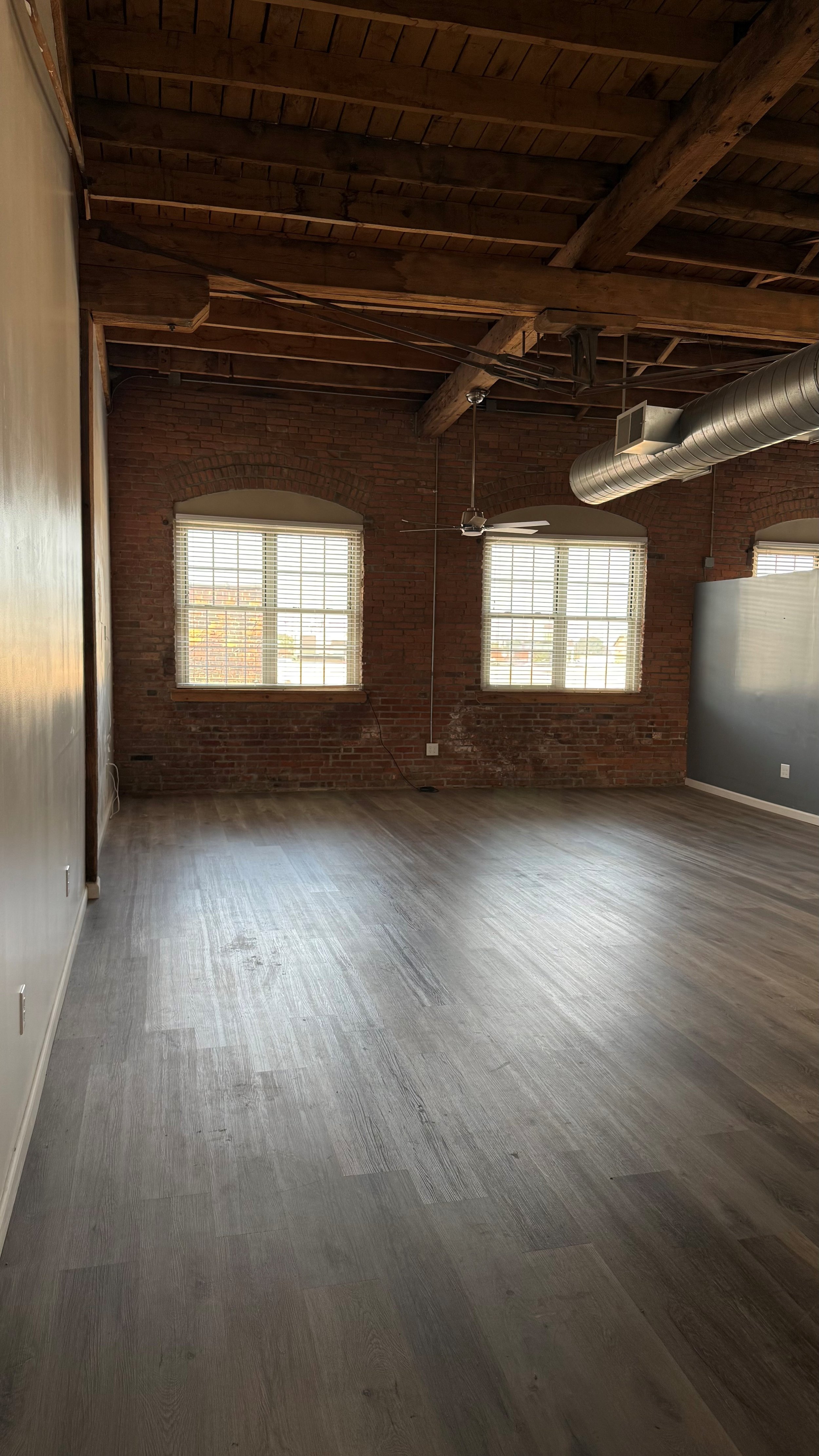 Empty loft-style room with wooden ceiling beams, brick walls, large windows with blinds, gray wood flooring, and industrial ductwork.