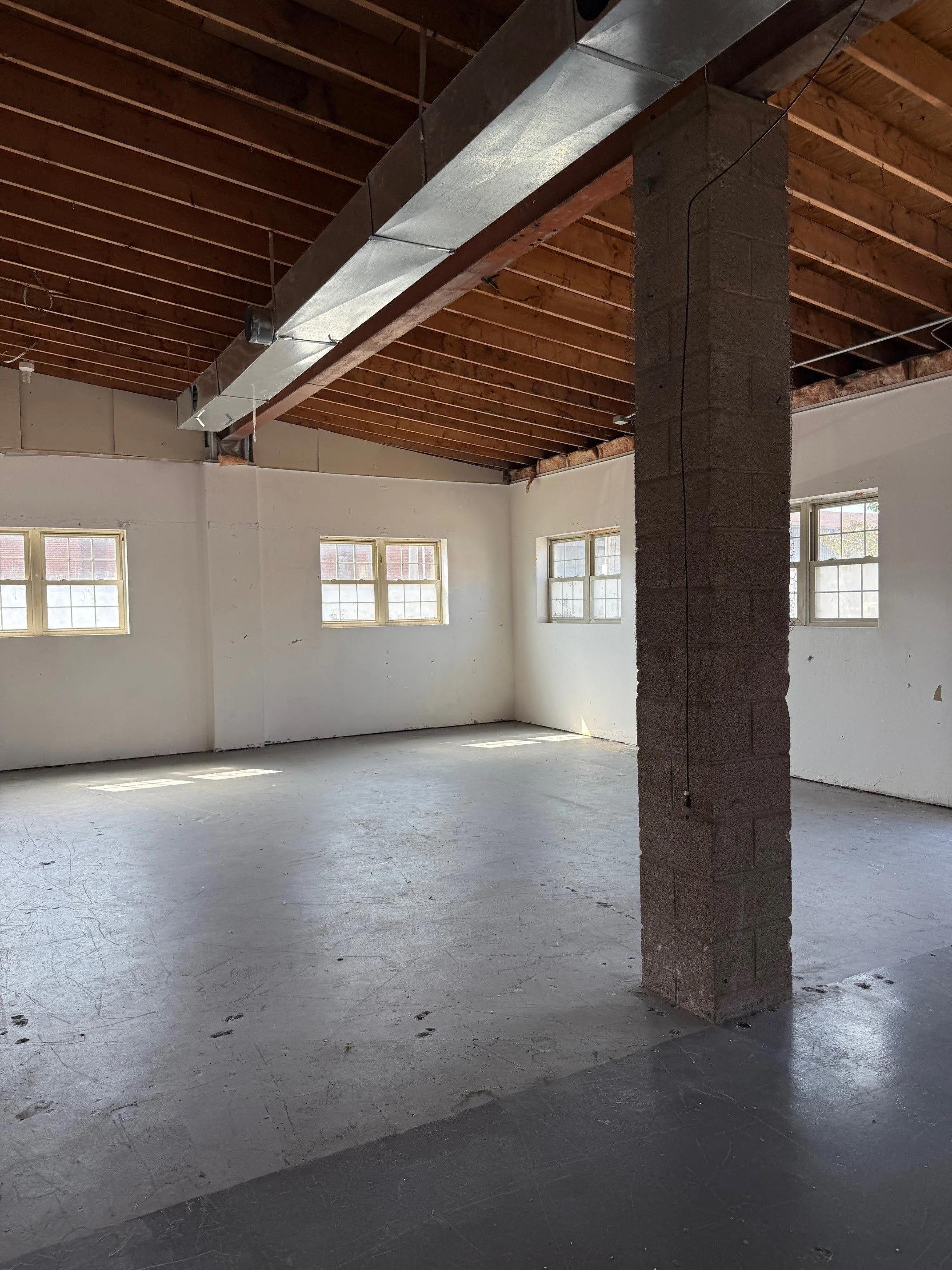 Empty room with white walls, concrete floor, four small windows, exposed wooden ceiling, metal ductwork, and a concrete pillar in the center.