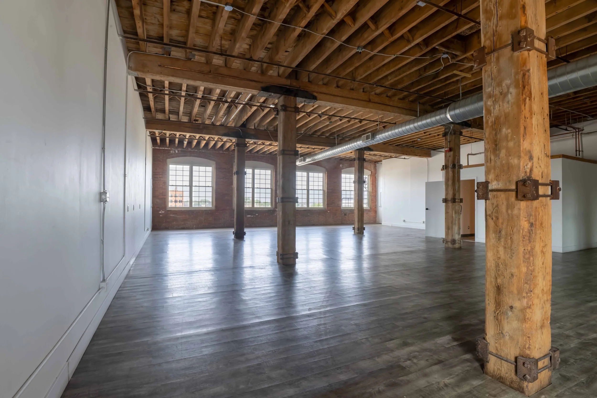 Large open space with exposed wooden ceiling beams, multiple large windows, brick wall, industrial-style support columns, polished wooden floor, and a central ventilation duct.