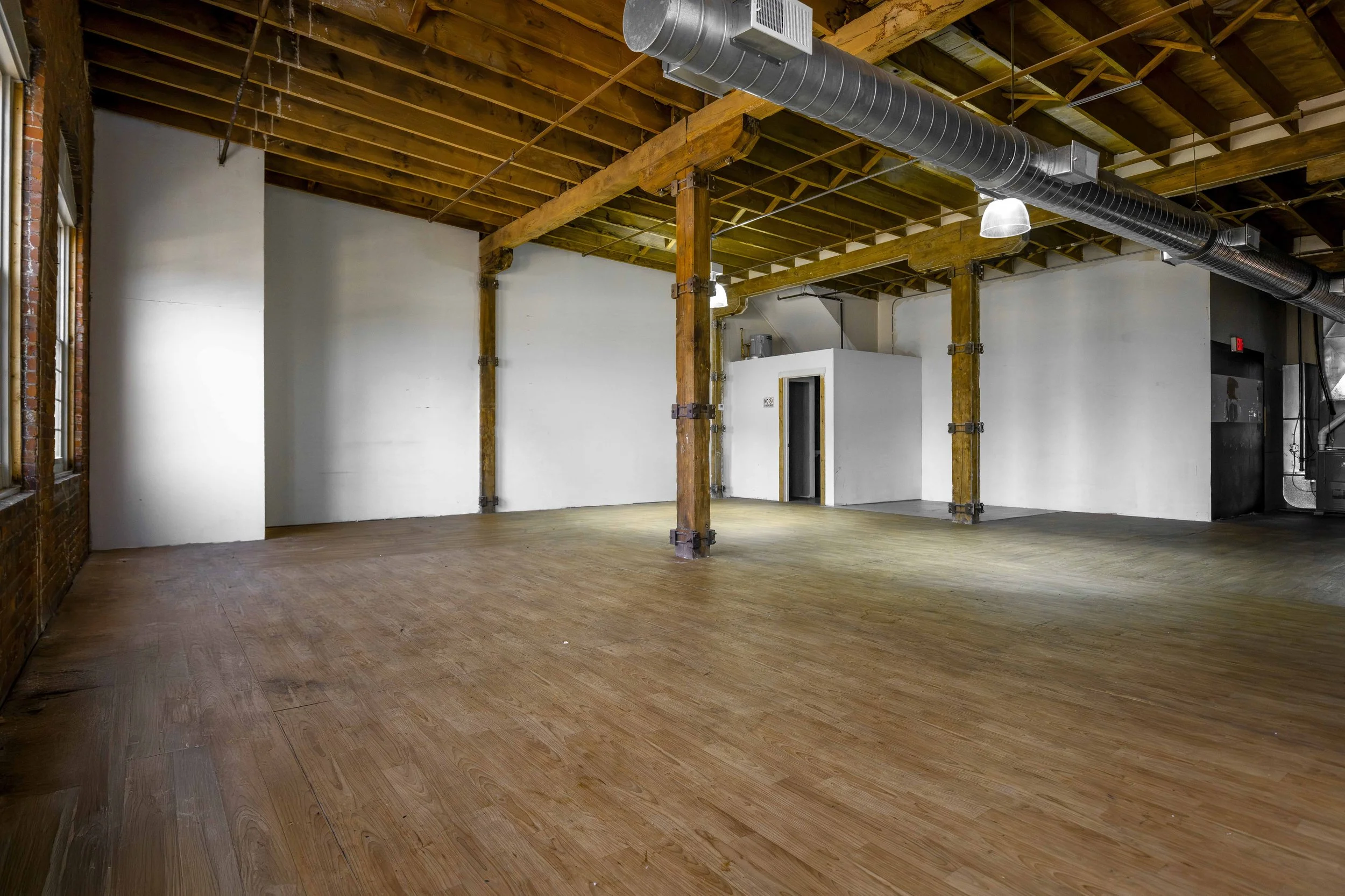 Empty room with wooden ceilings and floors, exposed beams, ducts, and white walls, with a small enclosed white structure and a large window on the left.