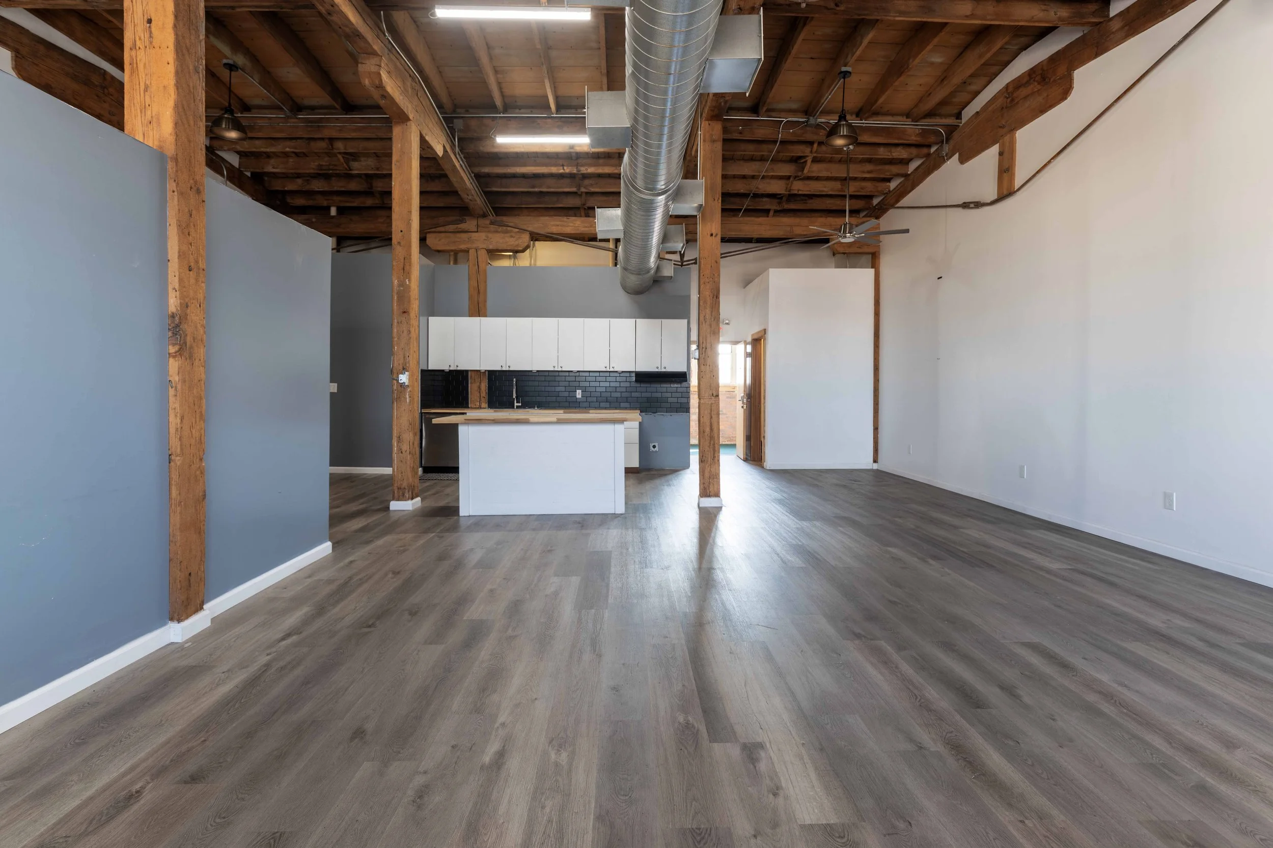 Empty open-concept loft with wooden ceiling beams, gray and white walls, exposed ductwork, and light gray wood flooring; a small kitchen area with white cabinetry and a black backsplash is visible in the background.