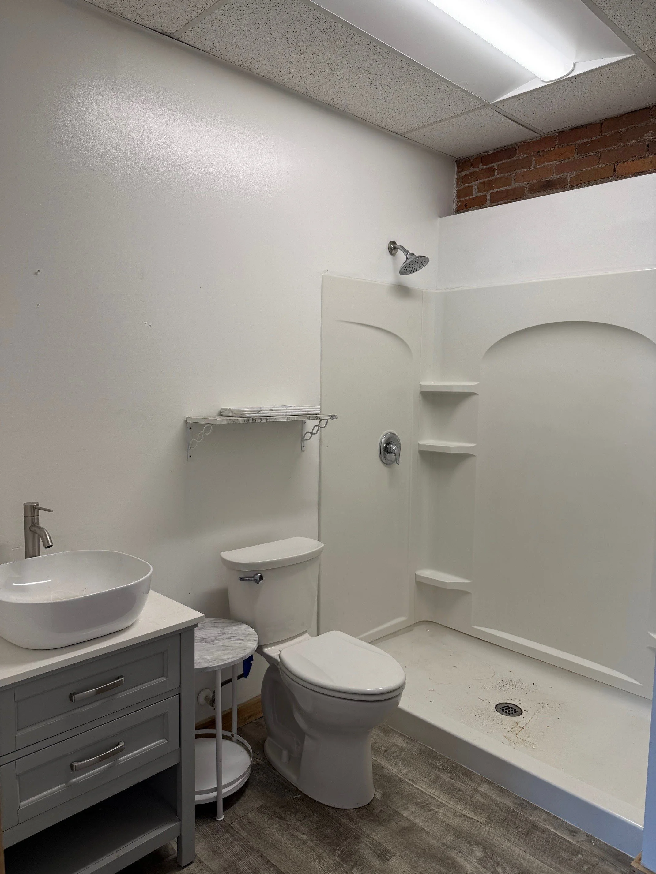 A small bathroom with a white toilet, a gray cabinet with a vessel sink, a small round side table, a wall-mounted shelf, and a walk-in shower with built-in shelves. The walls are mostly white with a brick section near the ceiling, and the floor is wo
