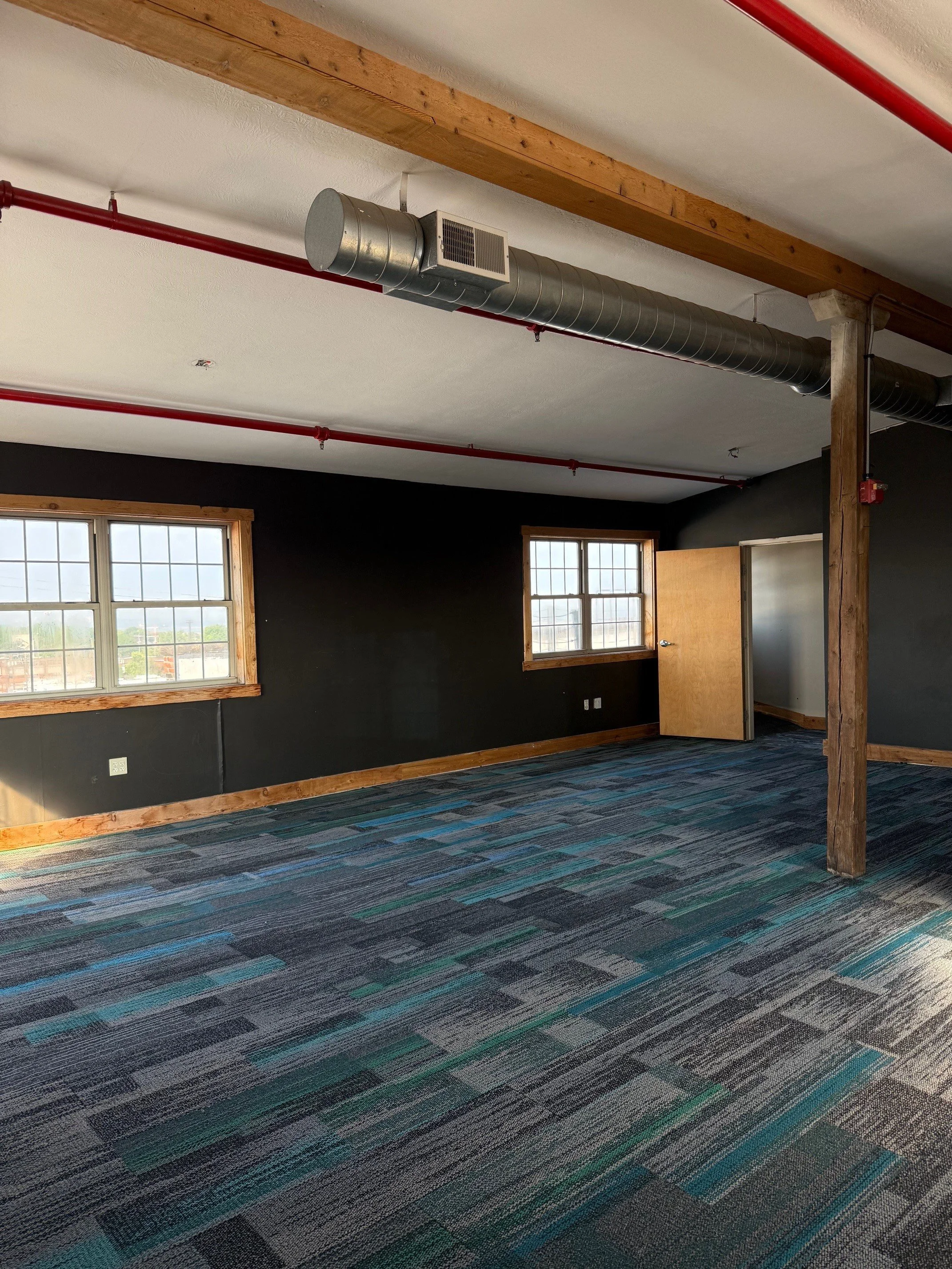 Empty room with two windows, black walls, wooden trims, carpeted floor, and exposed ductwork and beams on the ceiling.