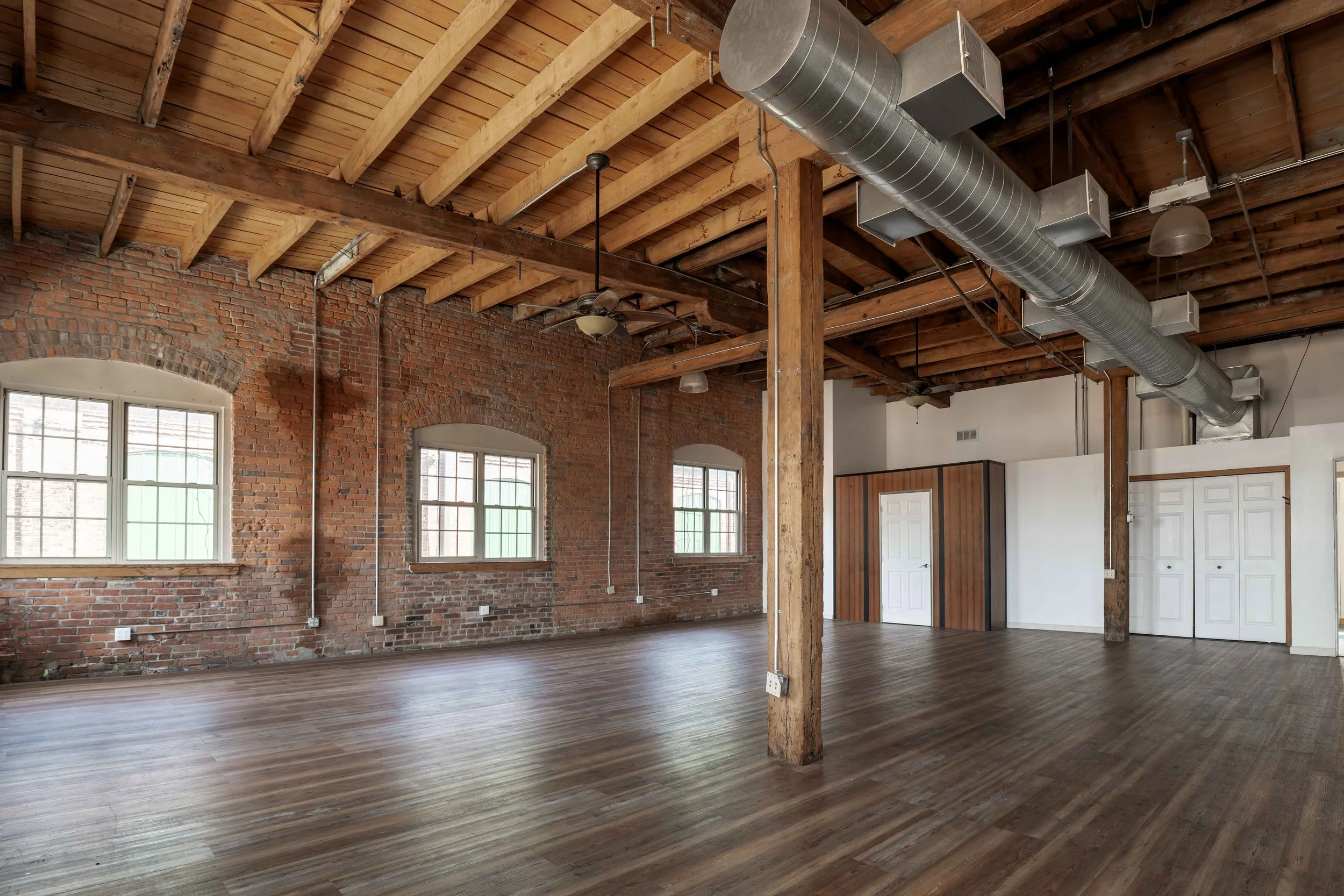 Interior of a spacious loft with exposed brick walls, wooden ceiling beams, large windows, and hardwood floors, featuring visible ductwork and built-in closet.
