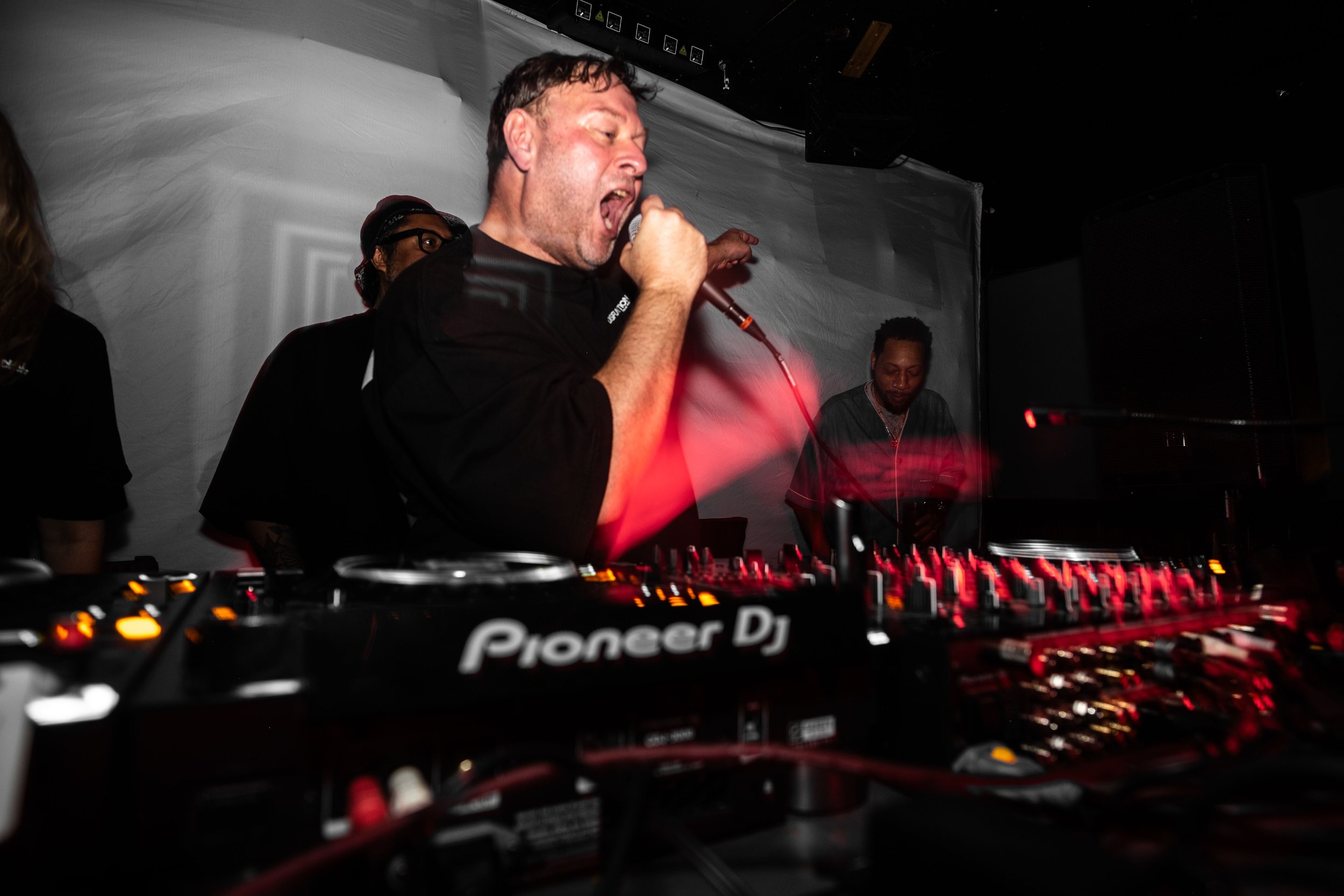 A DJ performs at a nightclub or event, singing into a microphone with a DJ console in the foreground and audience members in the background.