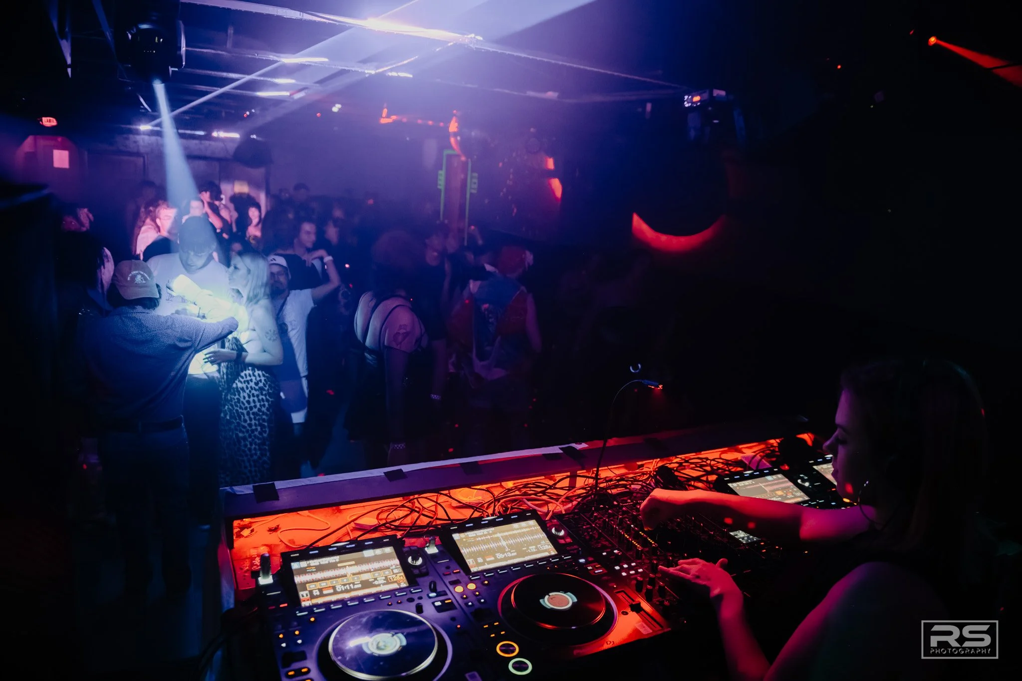 People dancing and socializing at a nightclub with a DJ playing music on a turntable setup, illuminated by colorful lights and laser beams.