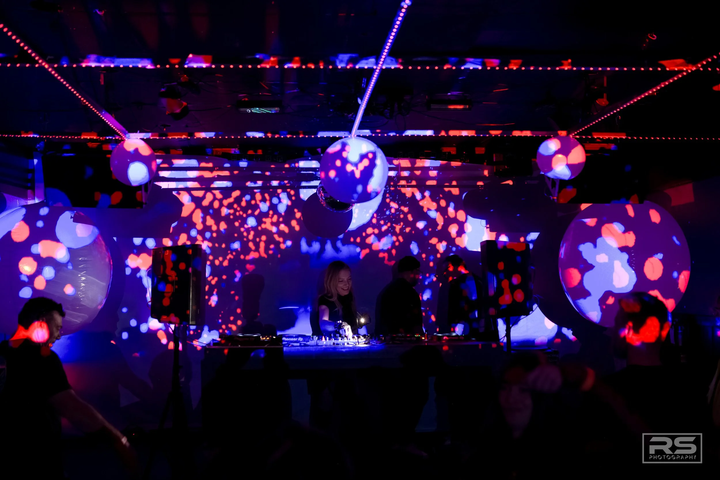 A nightclub scene with a DJ performing on stage, illuminated by colorful red, blue, and purple lights, with large illuminated spherical decorations and projected abstract patterns on walls and ceiling.