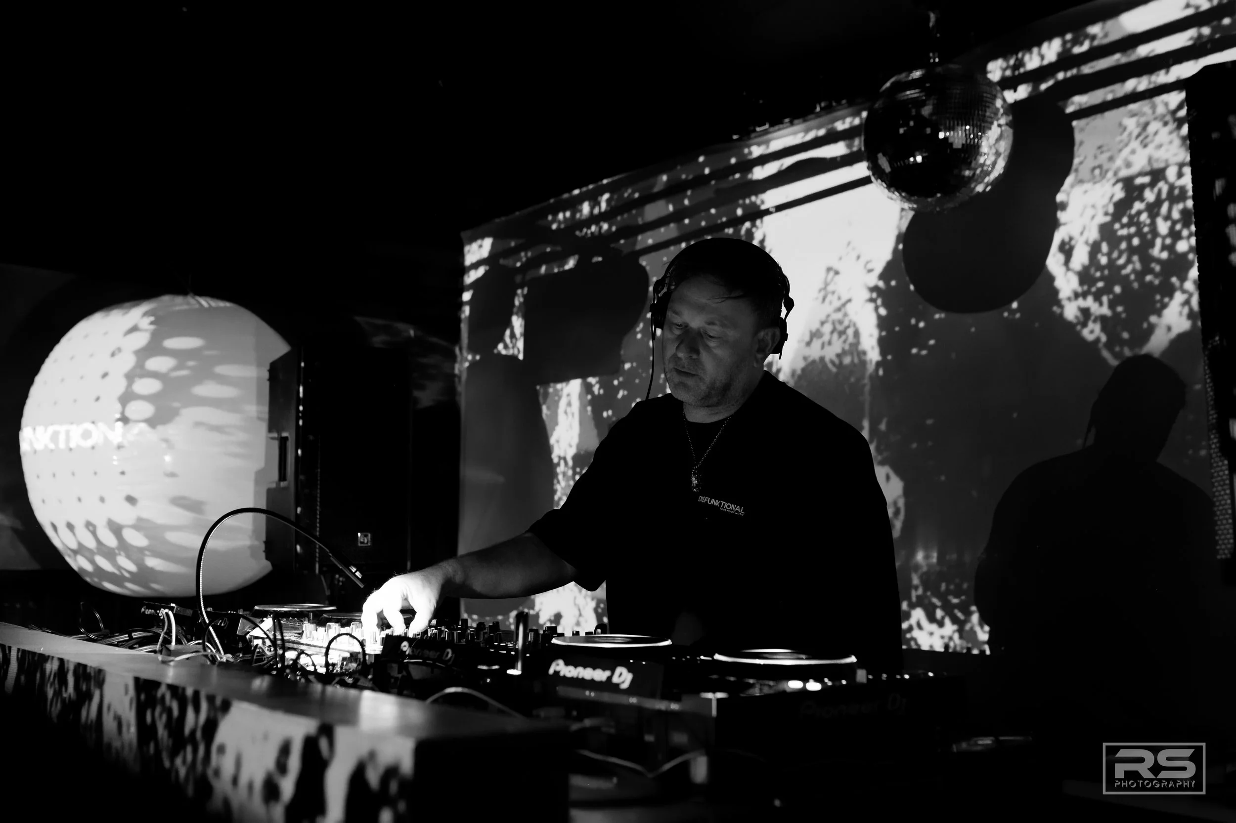 A DJ wearing headphones performs at a nightclub with a black and white visual display. The scene includes decorative lighting and a disco ball, creating an energetic atmosphere.